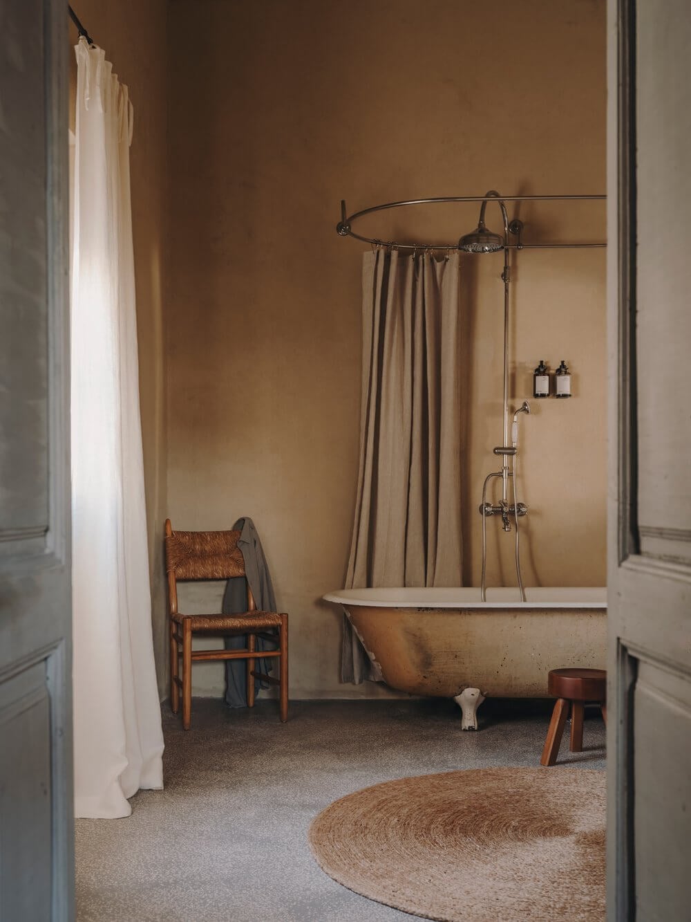 Casa Soleto, Europe — Minimalist bathroom with ochre walls, vintage clawfoot tub, brass rainfall showerhead, rush mat flooring, and rustic wooden chair.