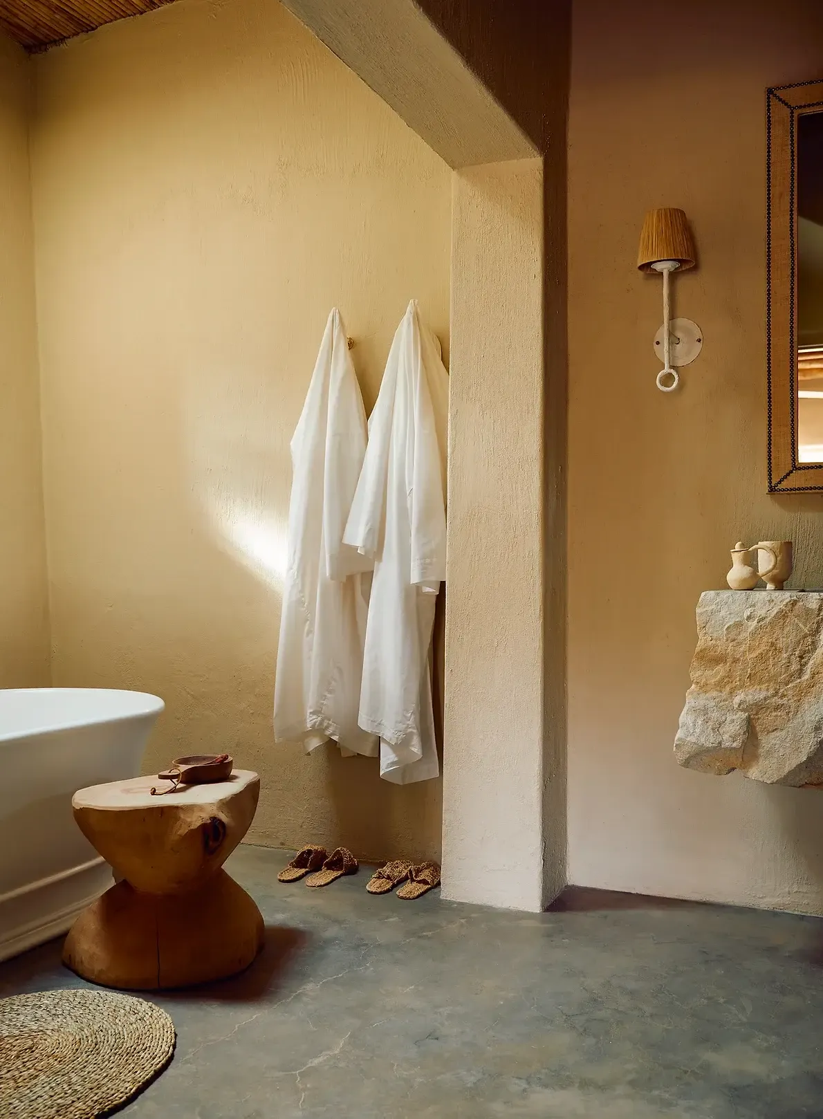 Sterrekopje, Africa — Minimalist bathroom with ochre walls, concrete floors, wooden vessel sink, white towels, and natural light casting geometric shadows.