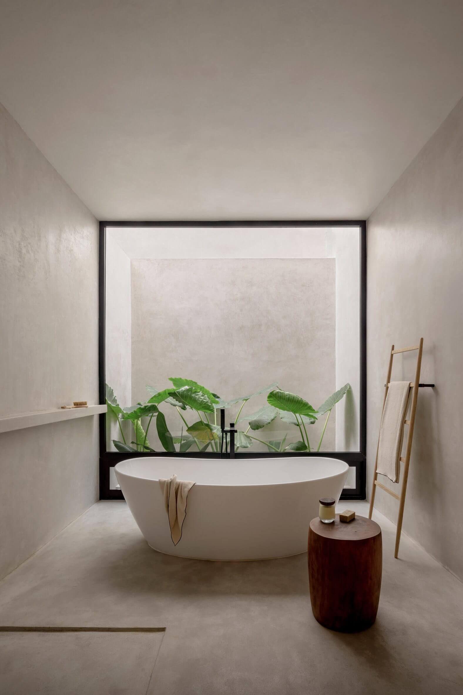 Villa Cava, Mexico — Minimalist bathroom with freestanding white oval bathtub, black-framed window overlooking green plants, wooden ladder towel rack, and polished concrete finishes.