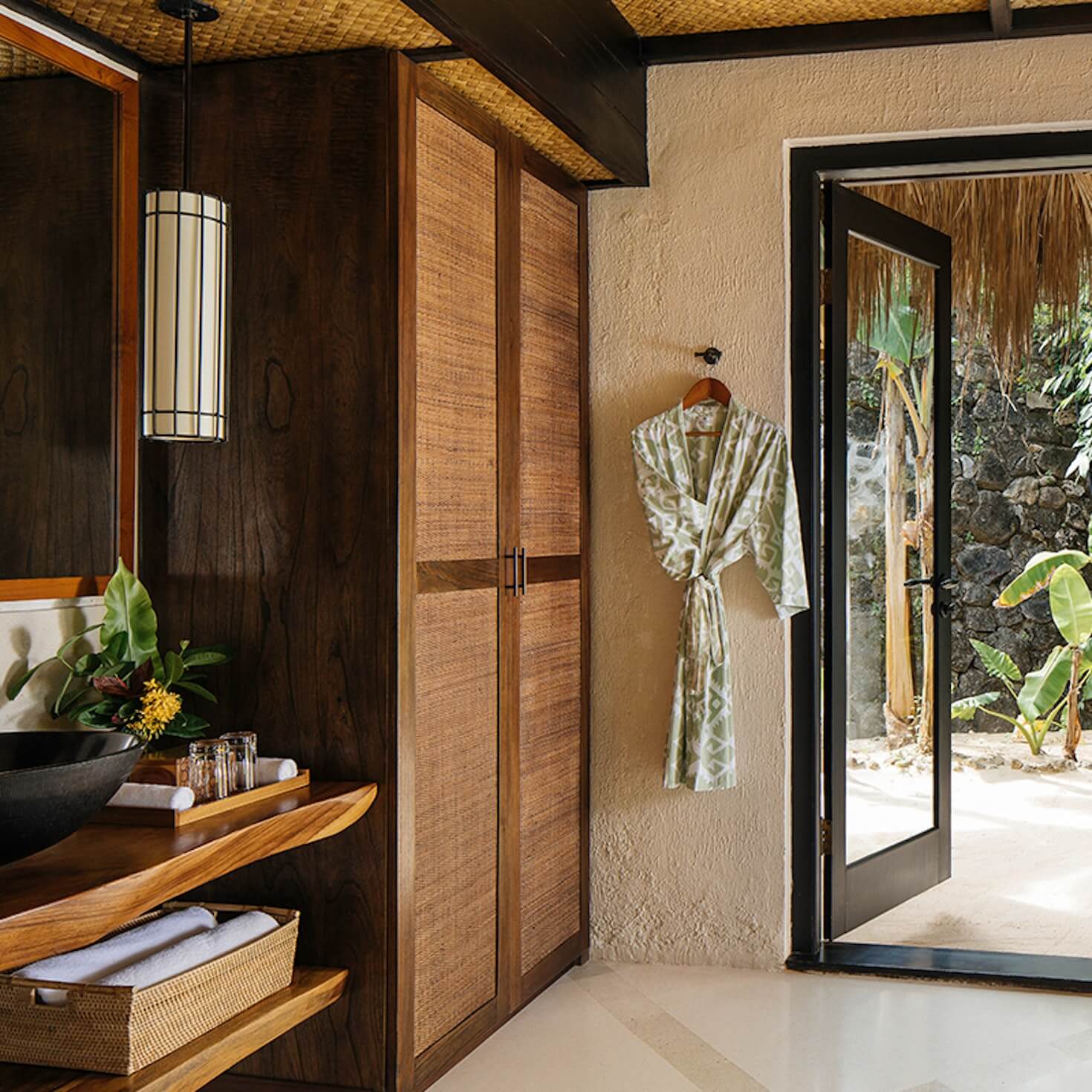NIHI Sumba, Asia — Spa bathroom with woven timber wardrobe, dark wood vanity, textured walls, pendant lighting, and garden views through glass doors.