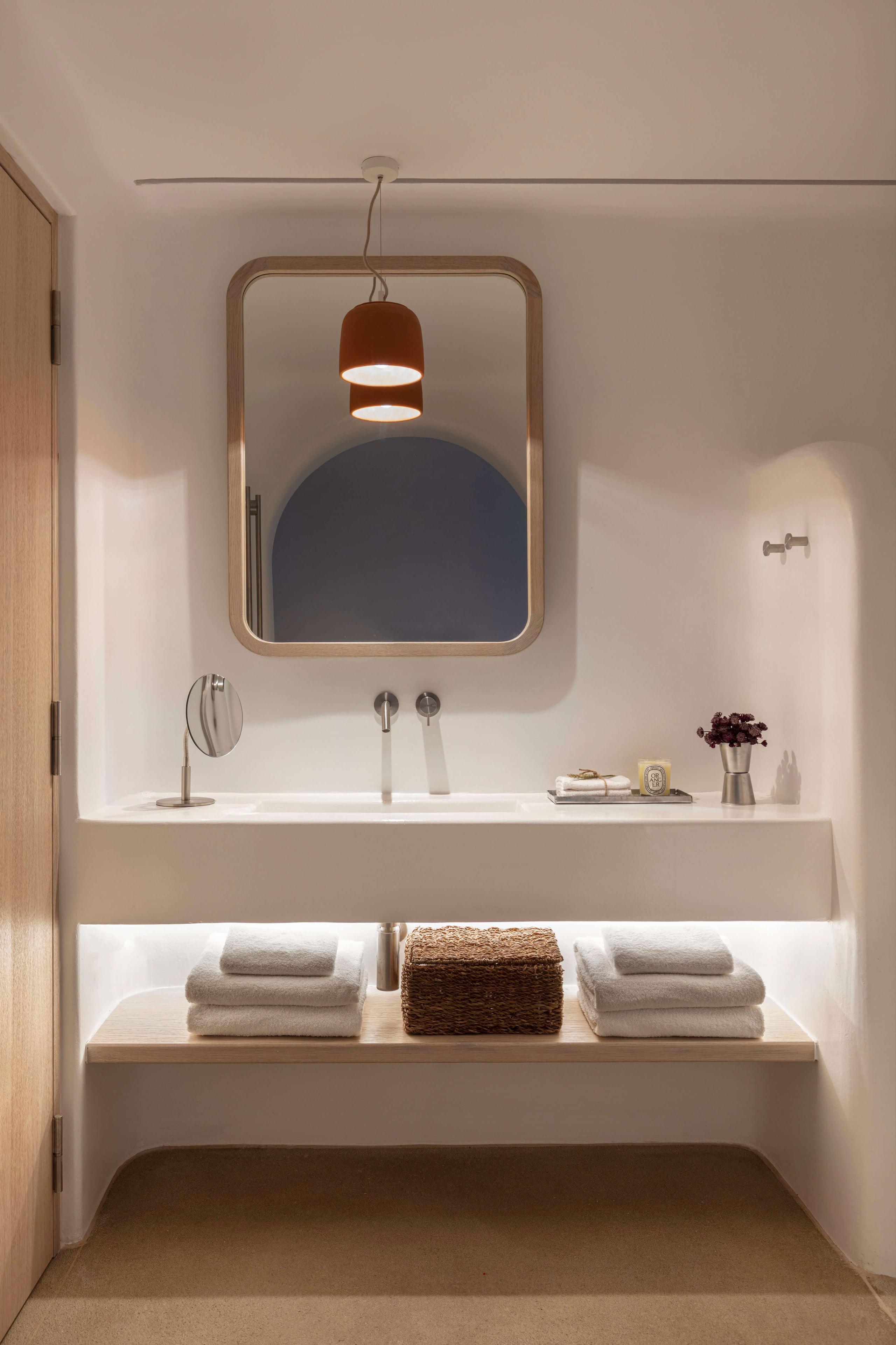 Villa Tama, Europe — Minimalist bathroom with rounded wood-framed mirror, terracotta pendant light, white vanity, and stacked linens on illuminated shelf.