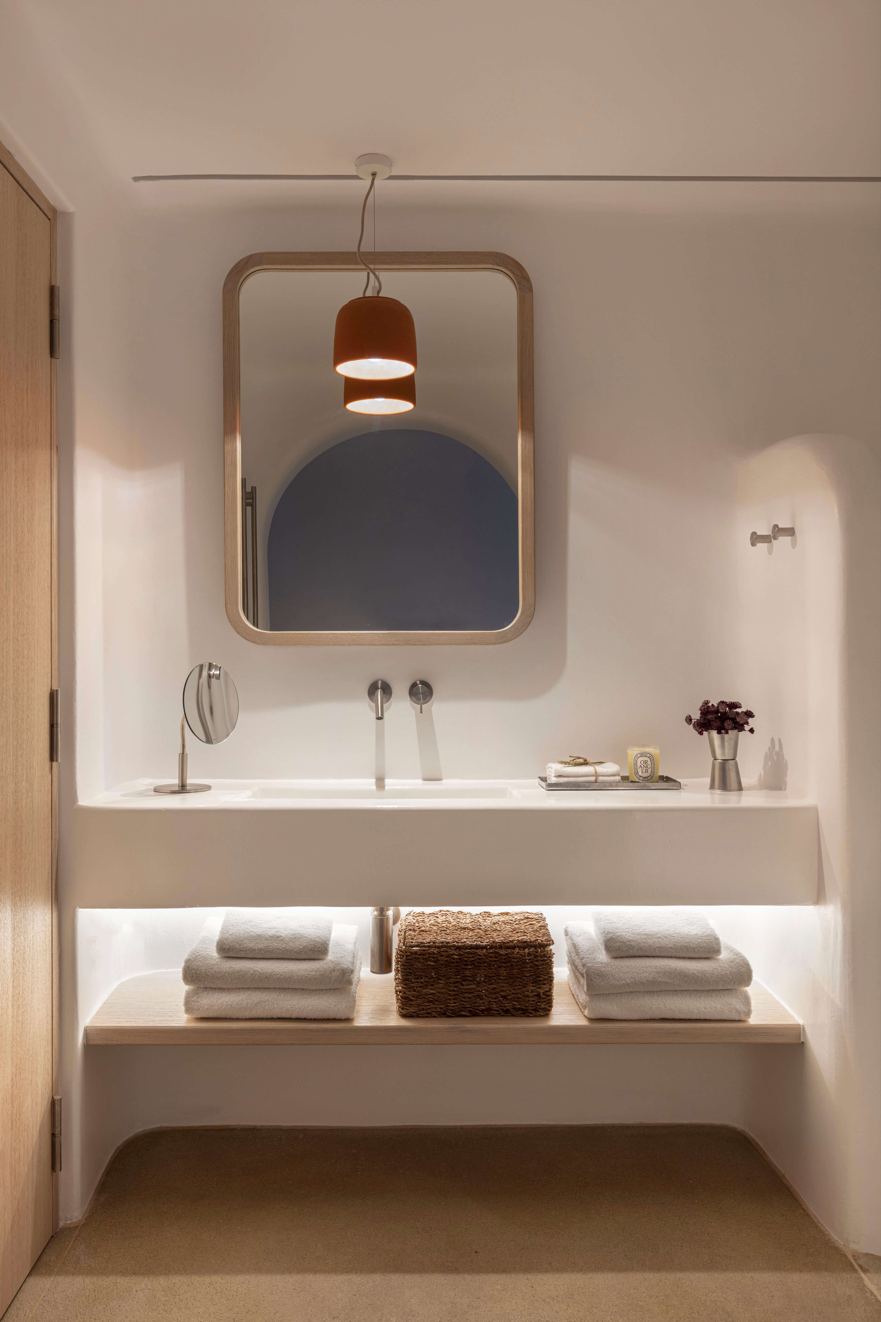 Villa Tama, Europe — Minimalist bathroom with rounded wood-framed mirror, terracotta pendant light, white vanity, and stacked linens on illuminated shelf.