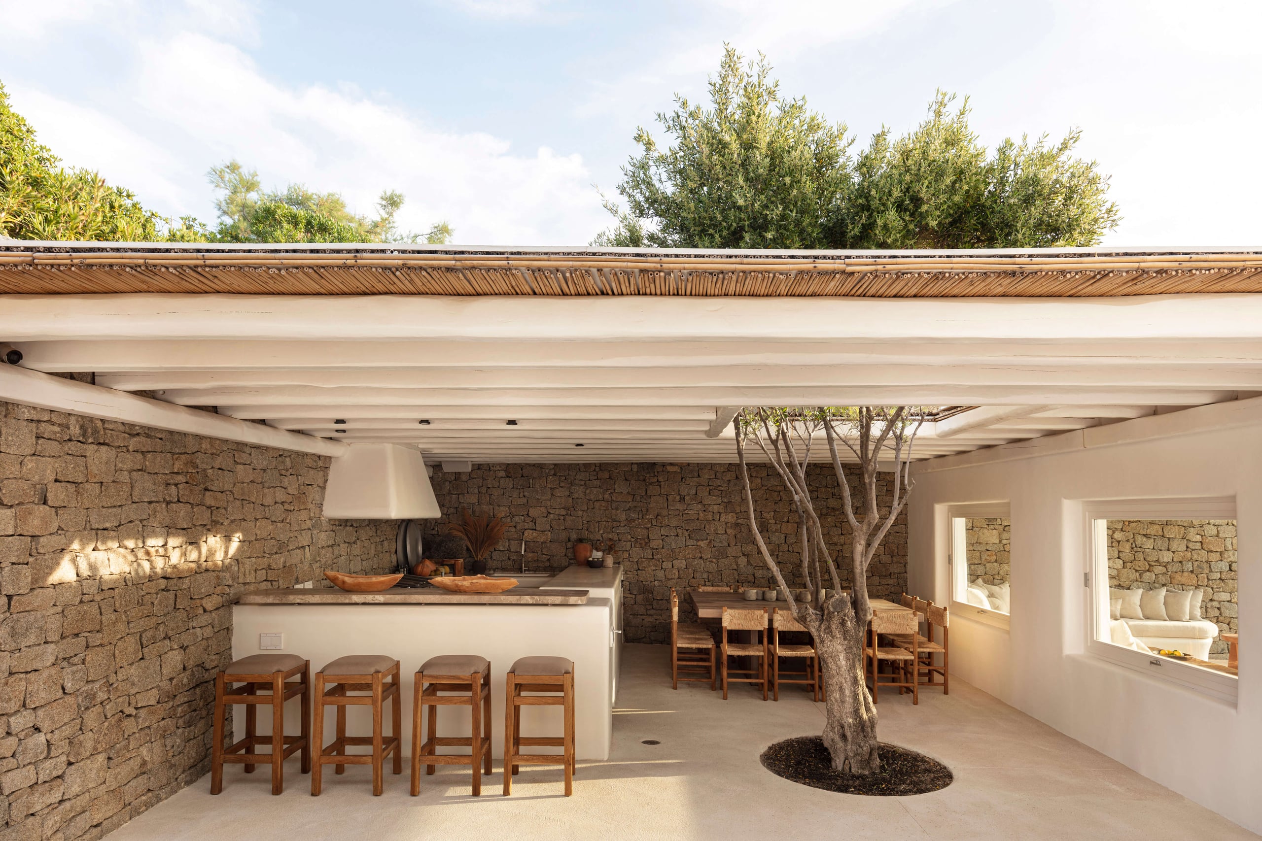 Villa Tama, Europe — Covered terrace with stone walls, minimalist kitchen island, wooden bar stools, dining area, and olive tree beneath geometric ceiling.