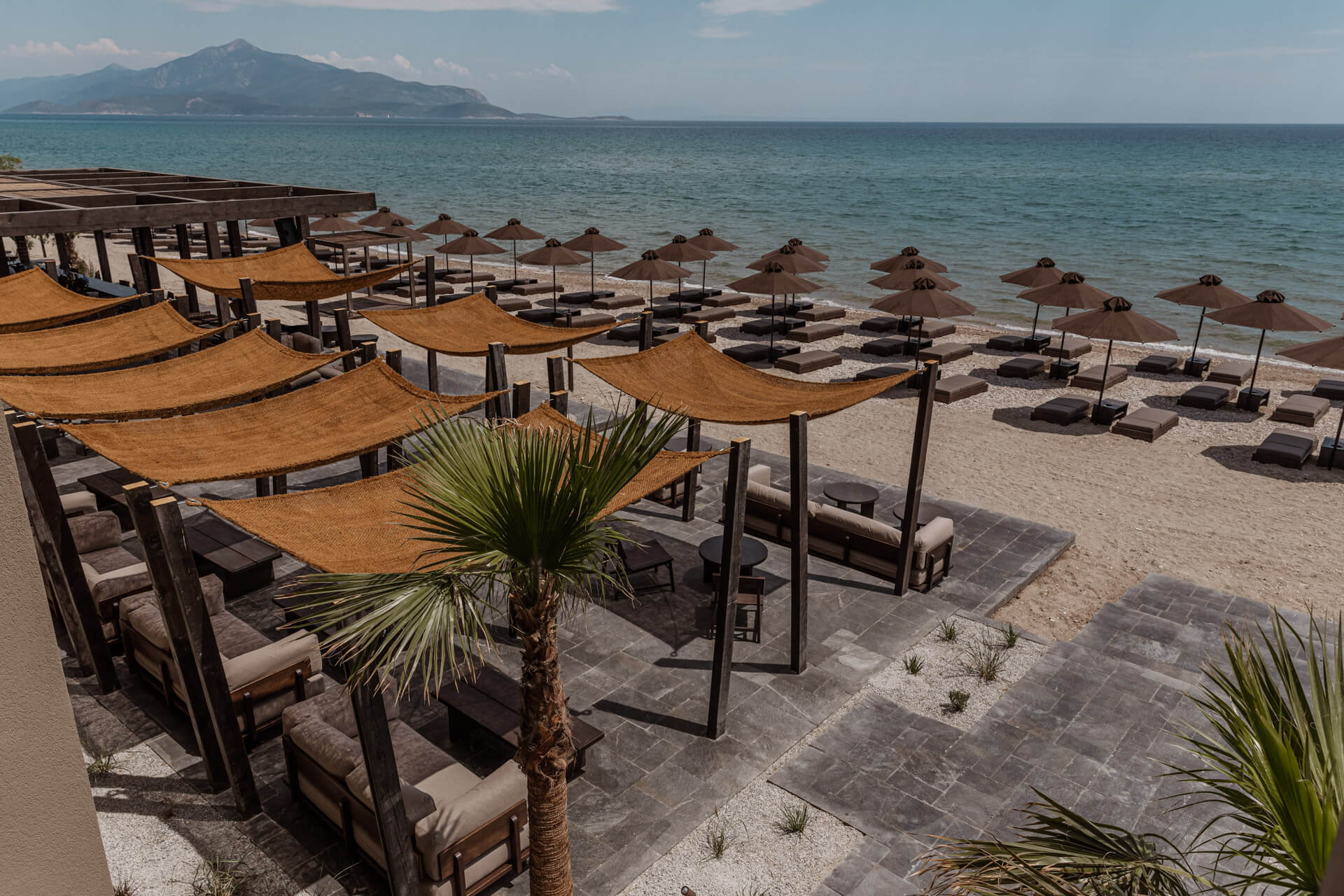 Beach Club, Europe — Ochre canvas canopies and loungers along pristine shoreline with mountain backdrop, luxury coastal hospitality design.