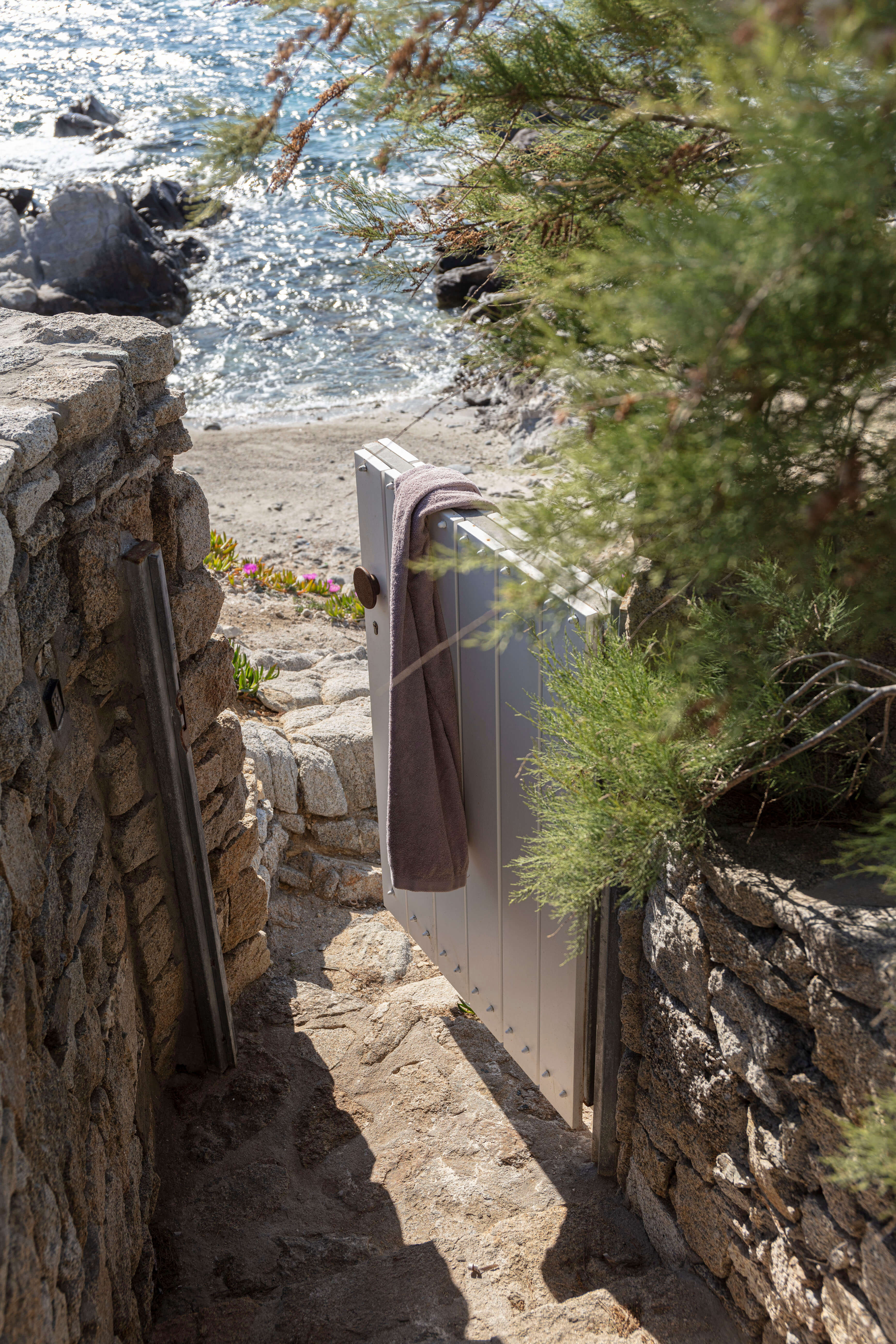 Villa Tama, Europe — Narrow stone pathway with wooden door opening toward Mediterranean beach, framed by weathered granite walls and pine branches.