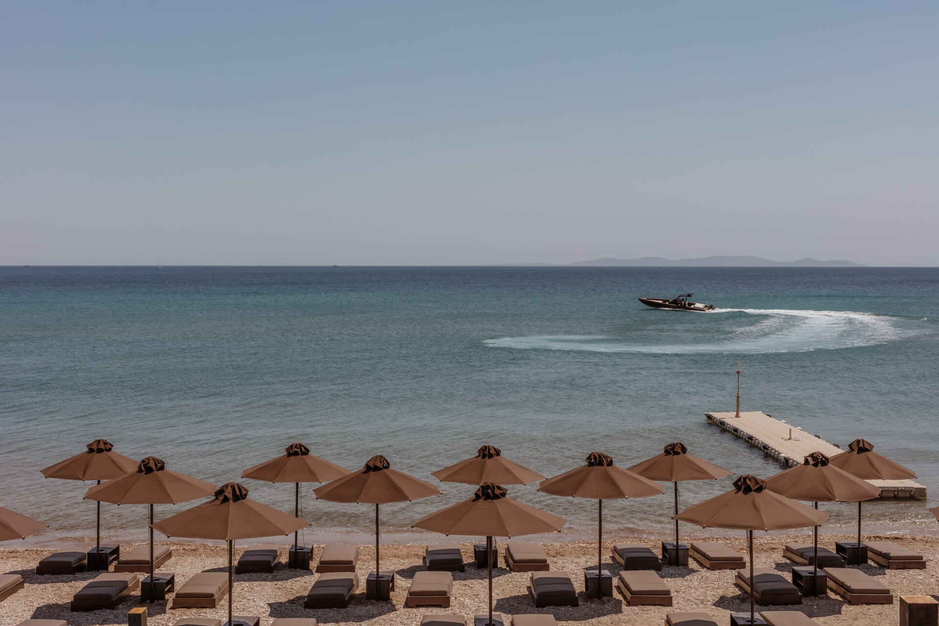 Casa Cook Samos, Greece — Taupe umbrellas and loungers line sandy beach with calm turquoise waters, speedboat, and pier under clear sky.