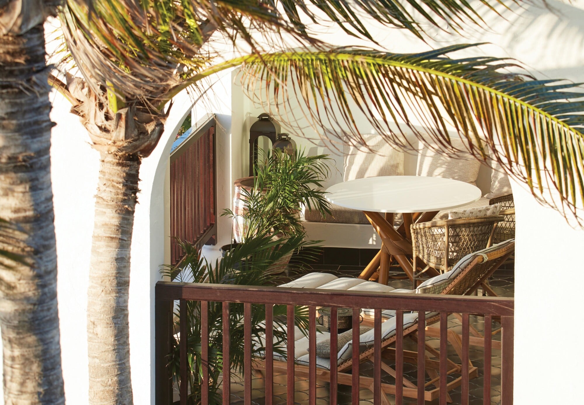 Cap Juluca, North America — White round table with wooden base on shaded terrace, woven chairs, potted palms, palm frond overhead, whitewashed architecture, coastal luxury design.