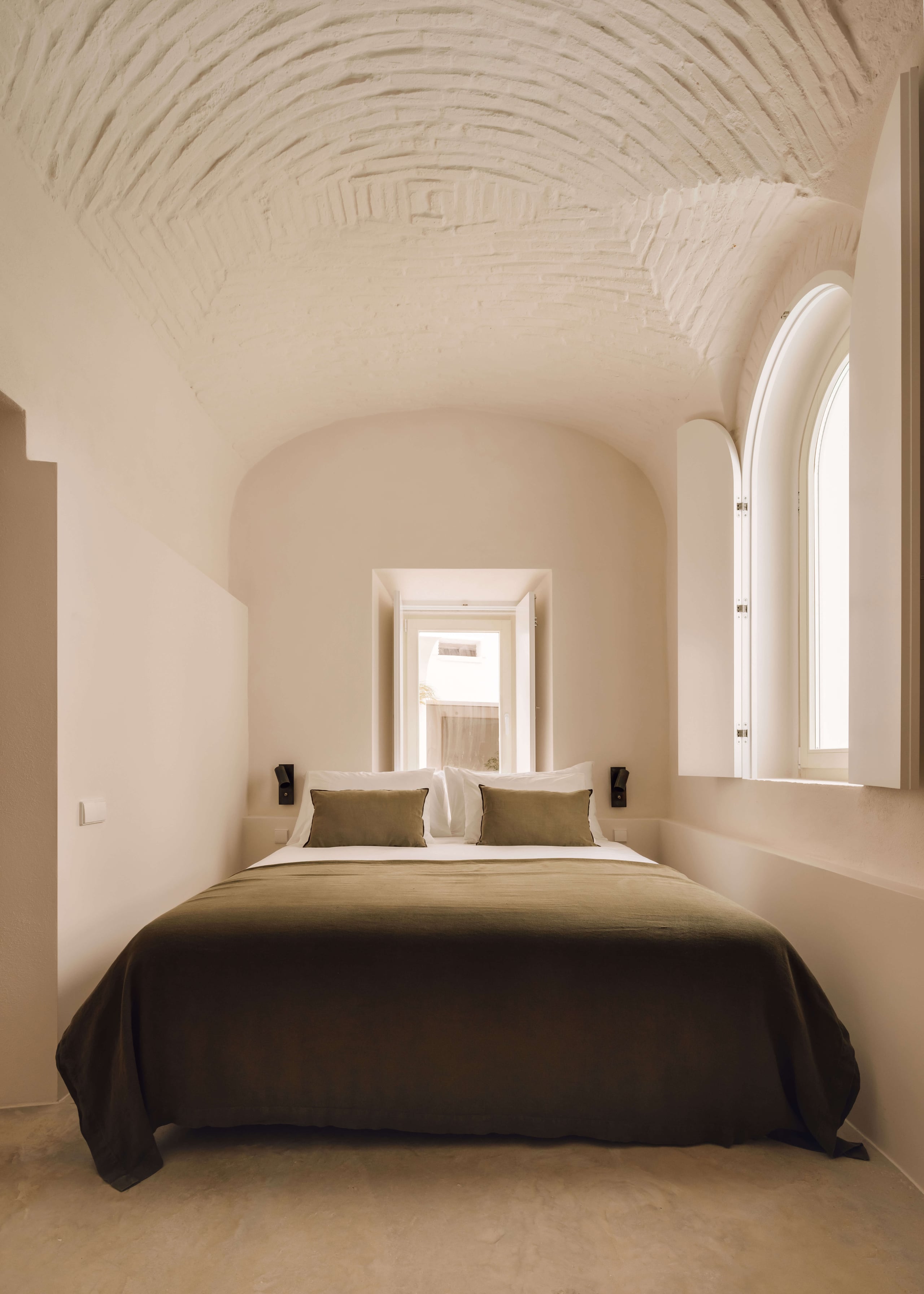 Casa Quatro, Europe — Minimalist bedroom with barrel-vaulted ceiling, arched alcove, dark bedcover, olive cushions, natural light from tall window, neutral plaster walls.