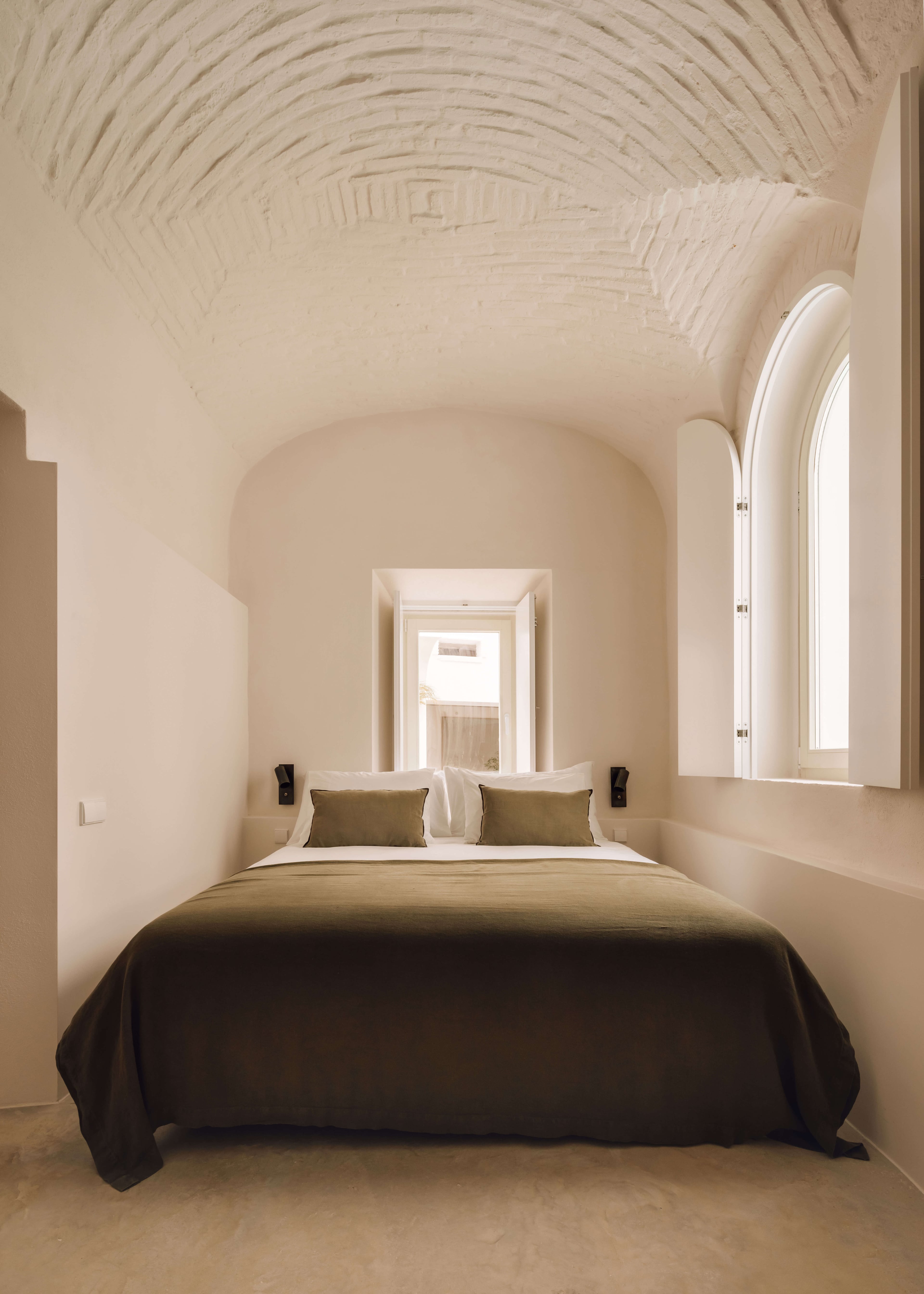 Casa Quatro, Europe — Minimalist bedroom with barrel-vaulted ceiling, arched alcove, dark bedcover, olive cushions, natural light from tall window, neutral plaster walls.