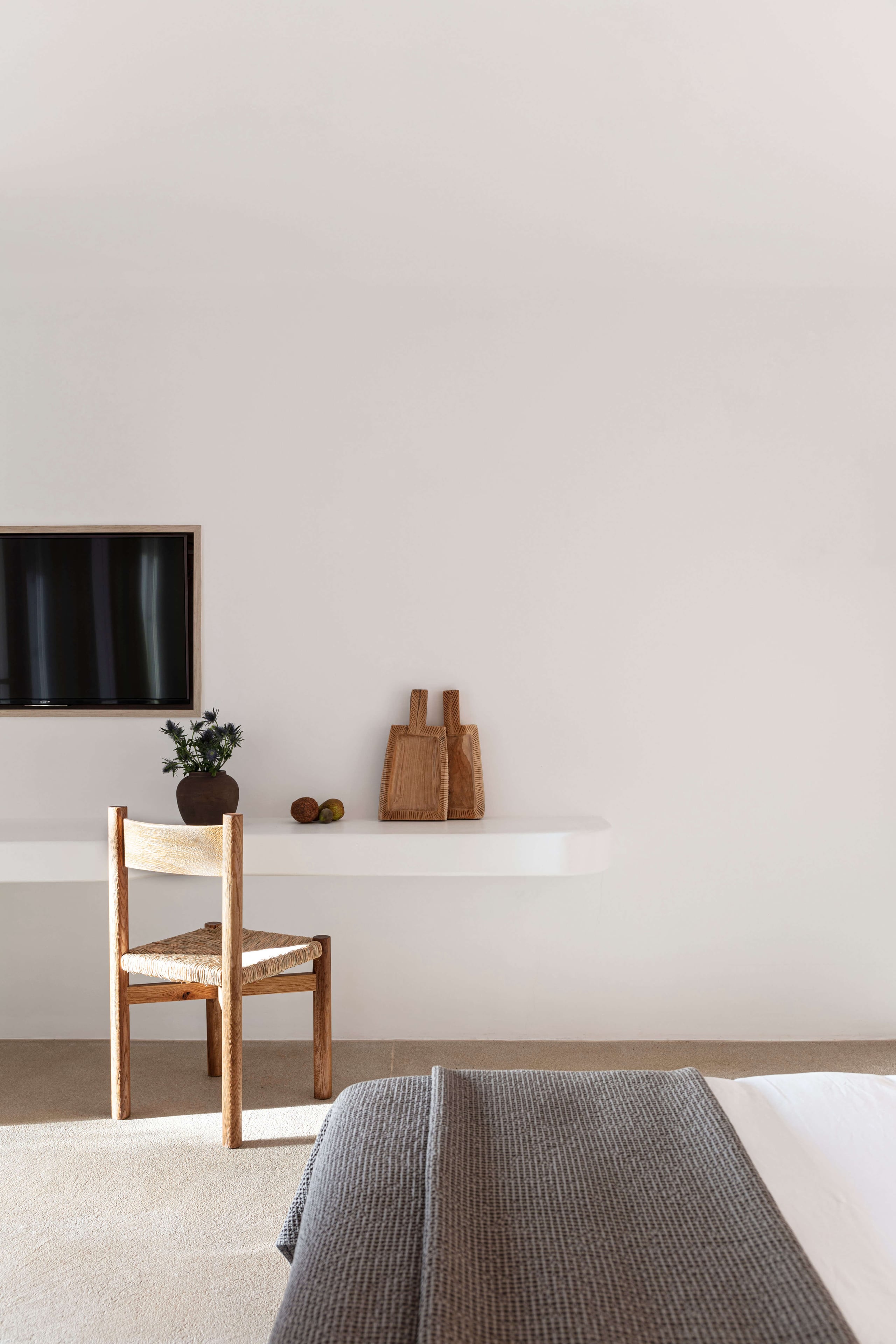 Villa Tama, Europe — Minimalist bedroom with woven chair, floating shelf, potted plant, wooden accessories, and gray bedding in natural light.