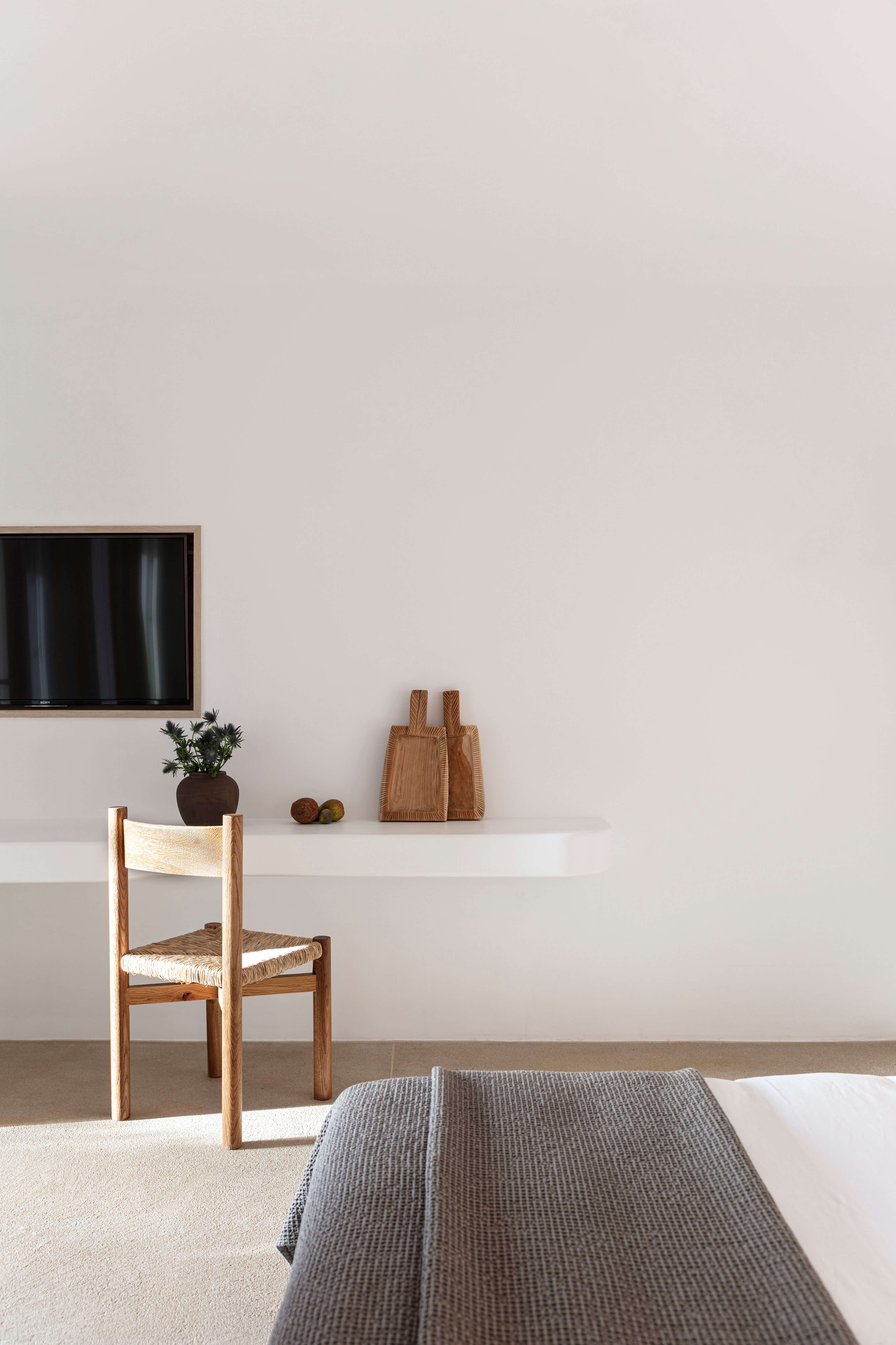 Villa Tama, Europe — Minimalist bedroom with woven chair, floating shelf, potted plant, wooden accessories, and gray bedding in natural light.