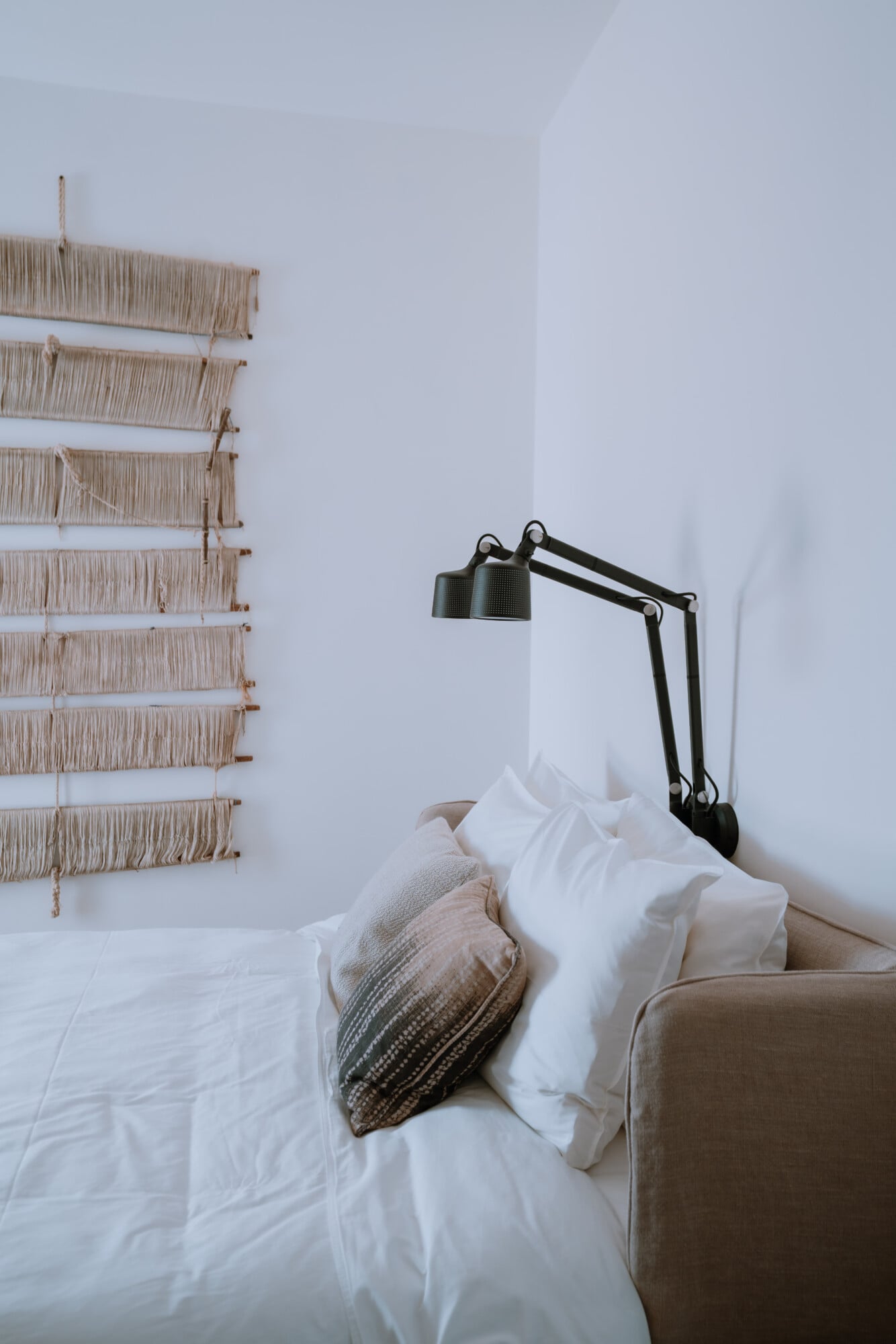 Villa Quercia, Italy — Minimalist bedroom with woven wall hanging, linen bedding, taupe pillow, and articulated black reading lamp against white walls.
