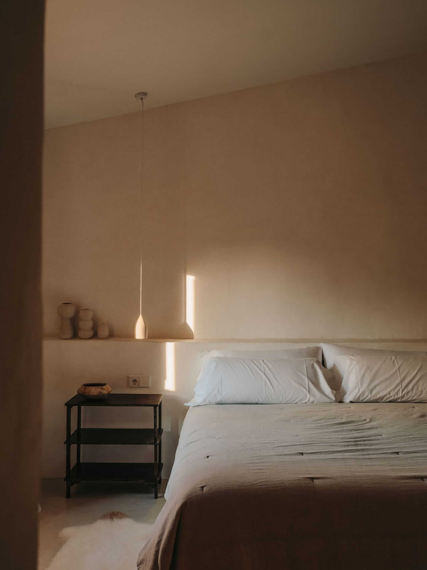 Can Lluïssó, Balearic Islands — Minimalist bedroom with pendant light, natural wood bed frame, black nightstand, and soft morning light through window.