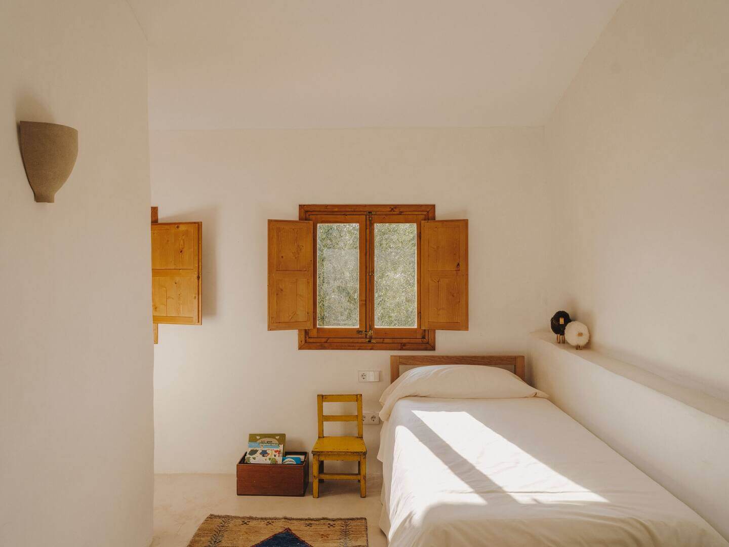 Can Lluïssó, Balearic Islands, Spain — Minimalist bedroom with wooden shutters, terracotta chair, white linens, and natural light flooding across stone floor.
