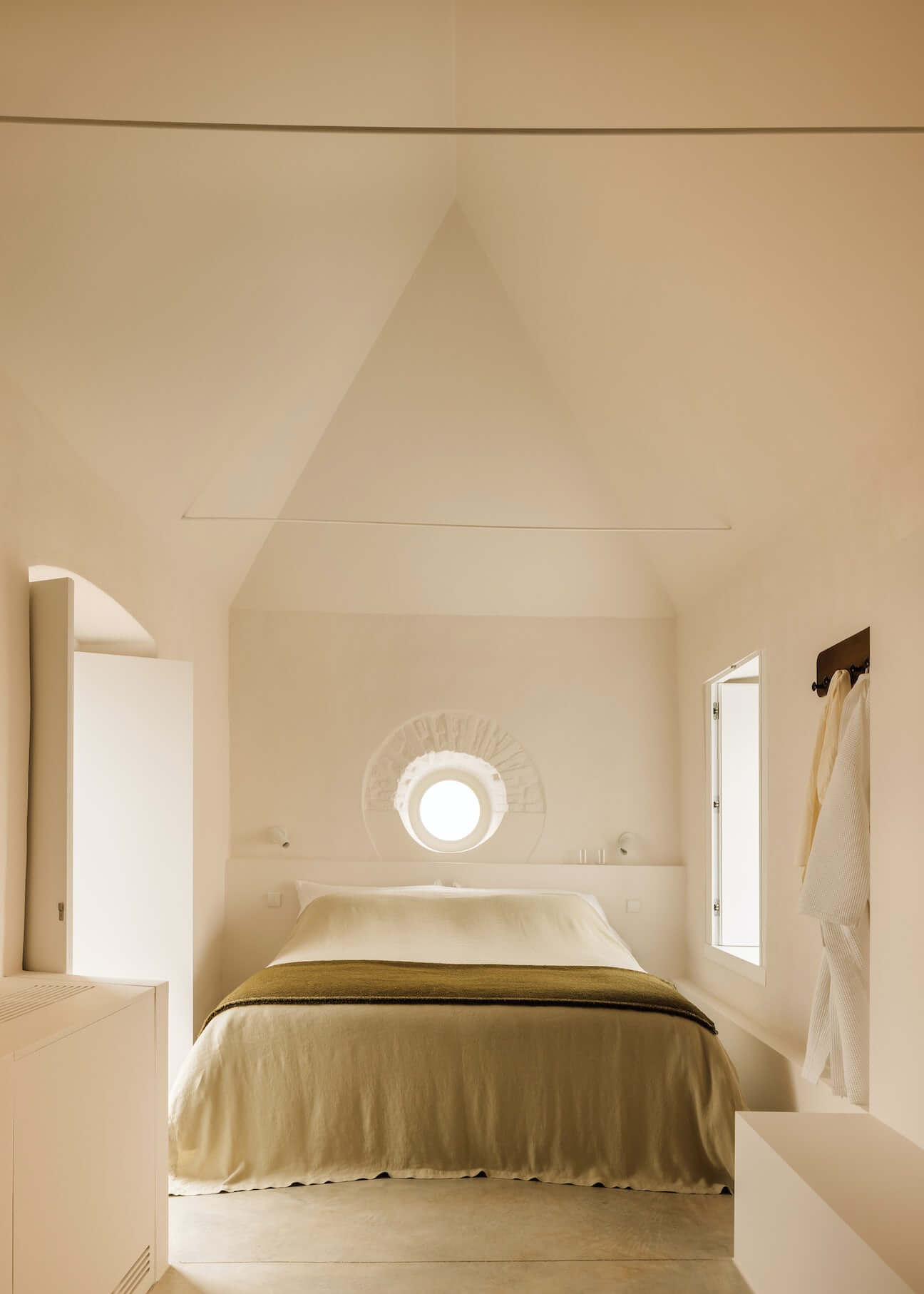 Casa Quatro, Europe — Minimalist bedroom with cream walls, pitched ceiling, circular window, olive bedcover, and natural light in luxury villa.