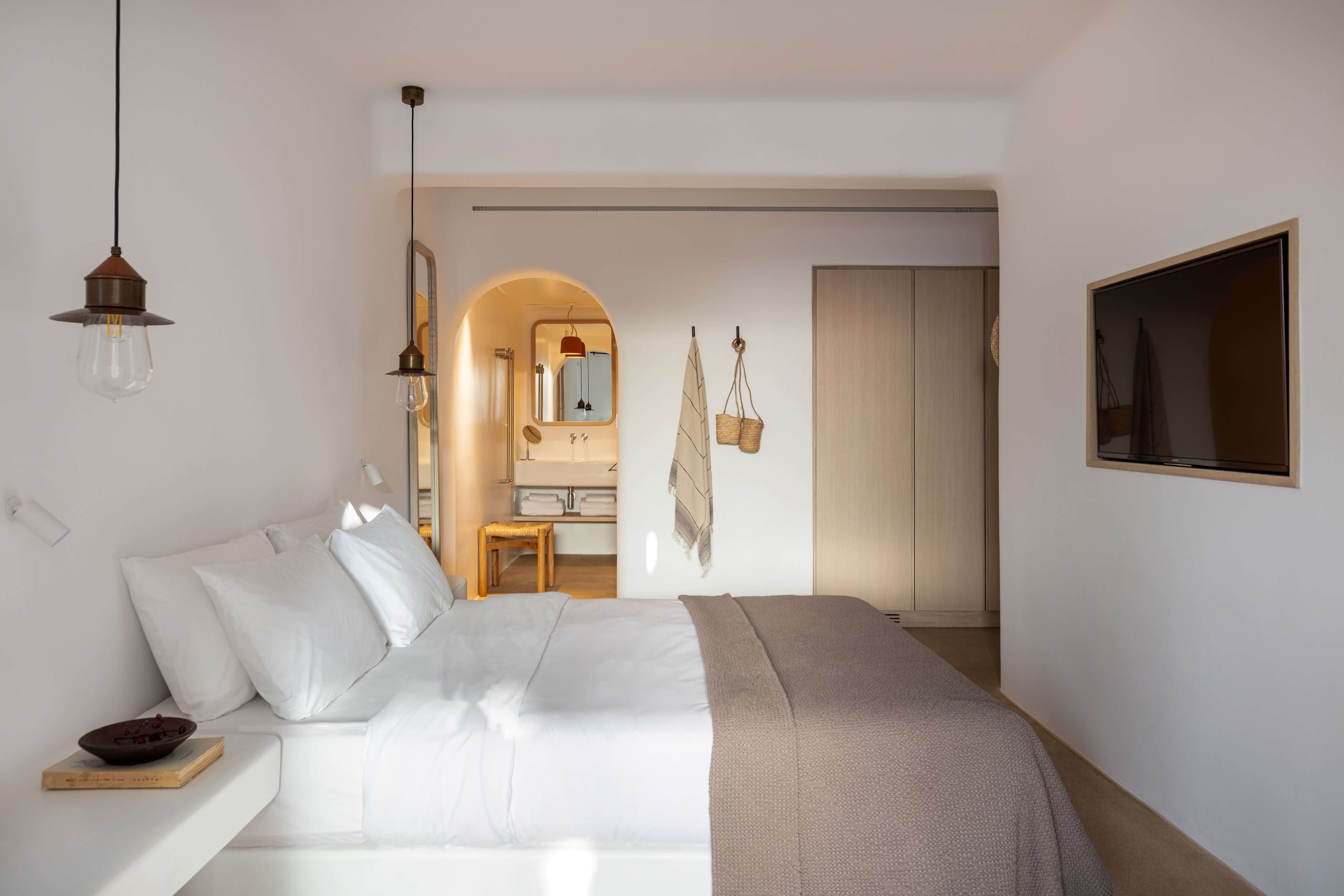 Villa Tama, Europe — Minimalist bedroom with ensuite bathroom featuring warm brass fixtures, arched mirror, white bedding, pendant lighting, and neutral palette throughout.
