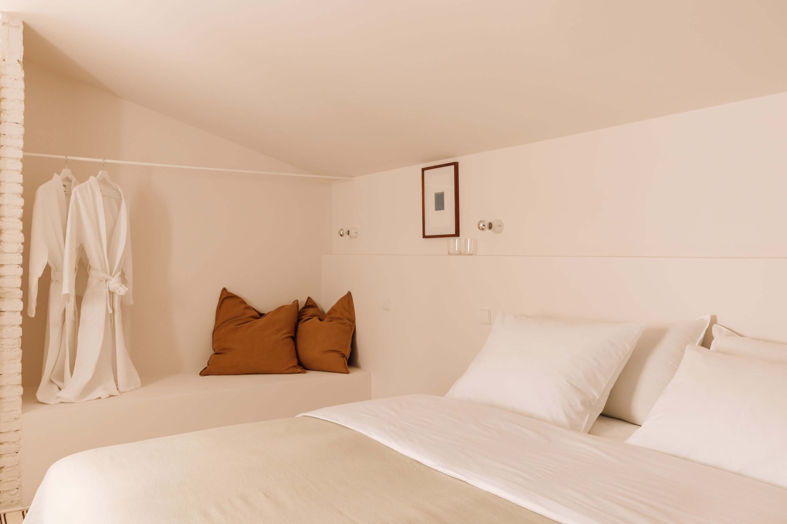 Casa Quatro, Europe — Minimalist bedroom alcove with built-in bench, terracotta pillows, white linens, cream walls, and natural light creating serene luxury interior design.