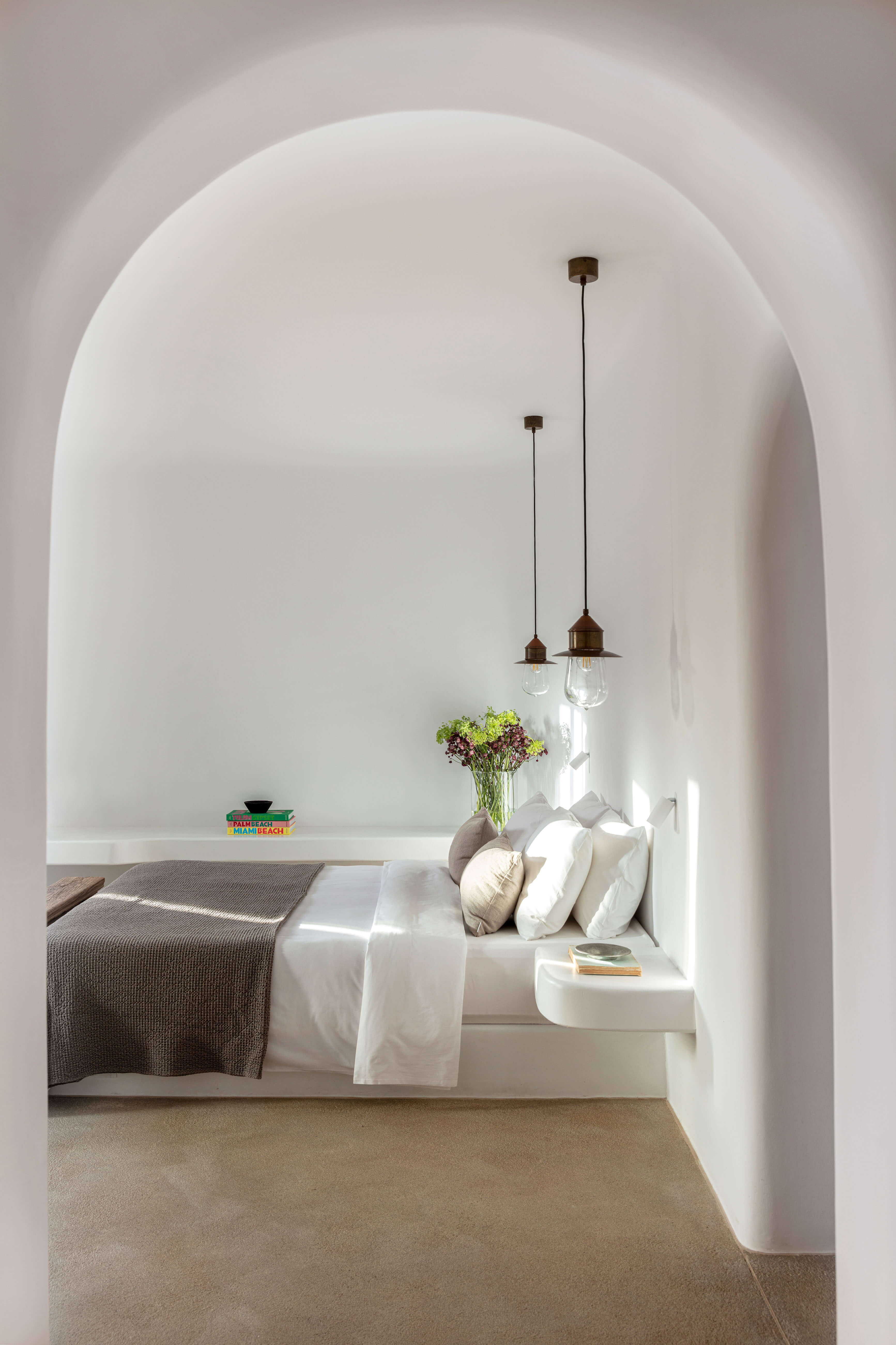 Villa Tama, Europe — Arched white bedroom alcove with built-in bed, linen pillows, pendant lights, fresh flowers, and minimalist Mediterranean design.