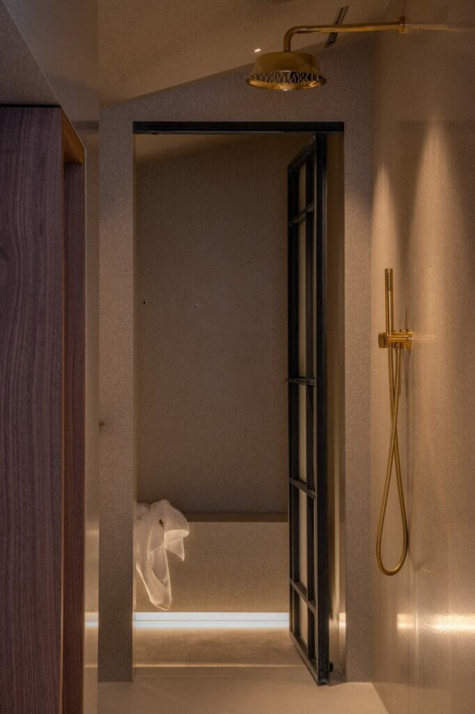 The Venice Venice Hotel, Europe — minimalist bathroom with brass rainfall showerhead, hand-held shower, black-framed glass partition, warm ambient lighting, neutral stone walls, and folded white towel.