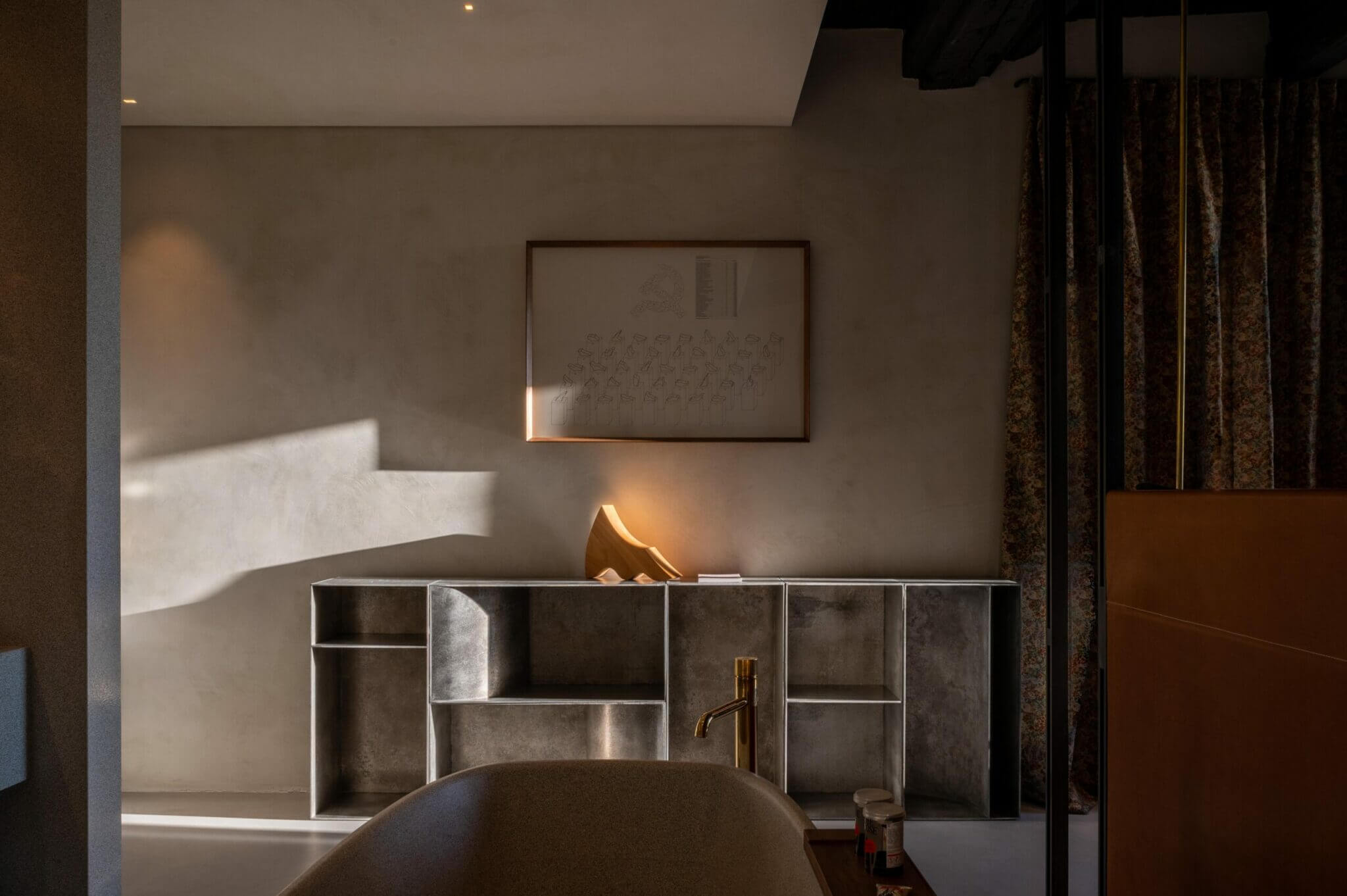 The Venice Venice Hotel, Europe — Minimalist bathroom with metallic storage unit, brass faucet, curved dark basin, framed artwork, and warm natural light casting geometric shadows on neutral walls.