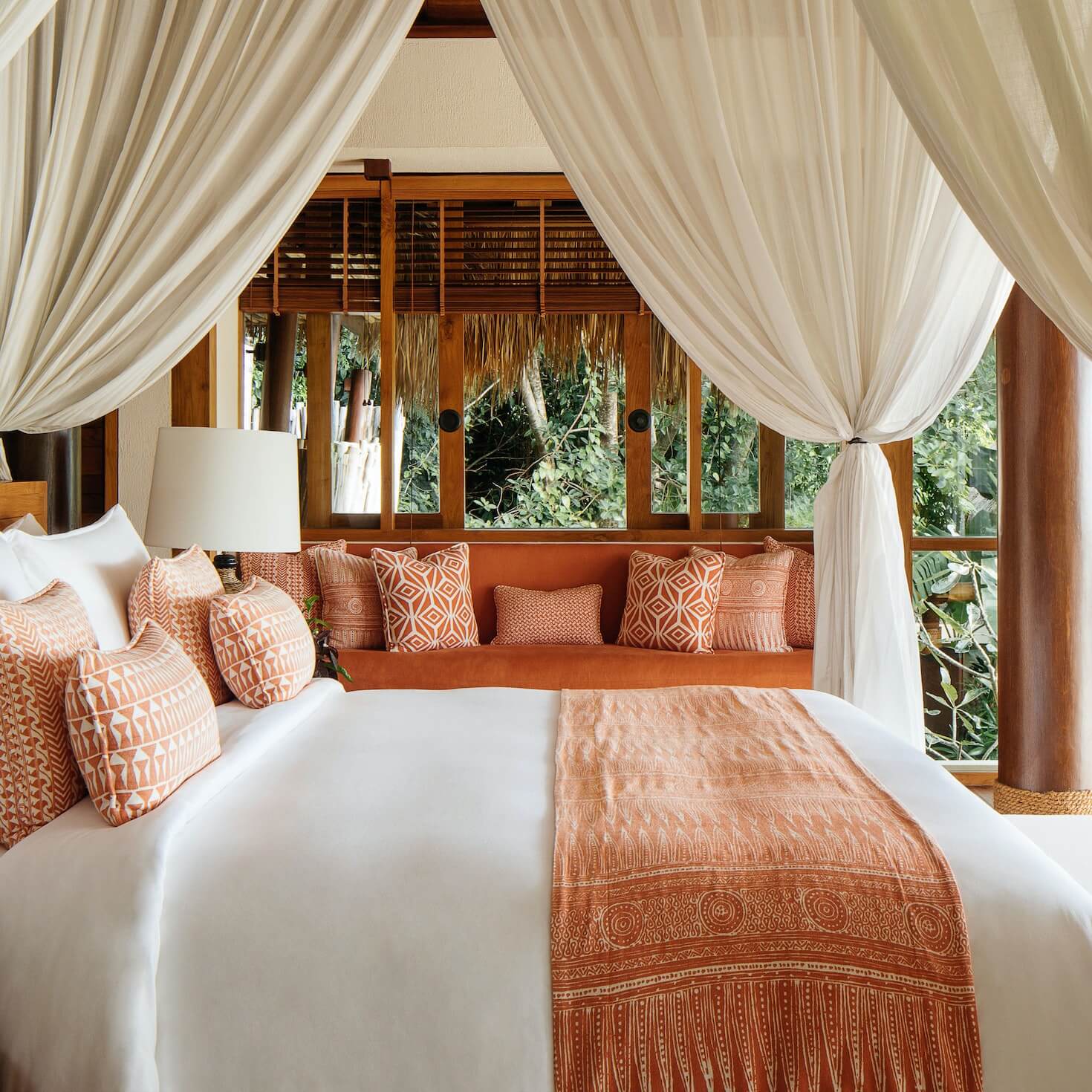 NIHI Sumba, Asia — Canopied bed with cream drapes, terracotta patterned pillows, wooden pavilion overlooking tropical garden, warm natural light.