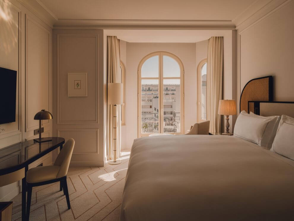 Carlton Cannes, Côte d'Azur, France — Sunlit bedroom with arched windows, herringbone floors, neutral palette, and cityscape views from balcony.