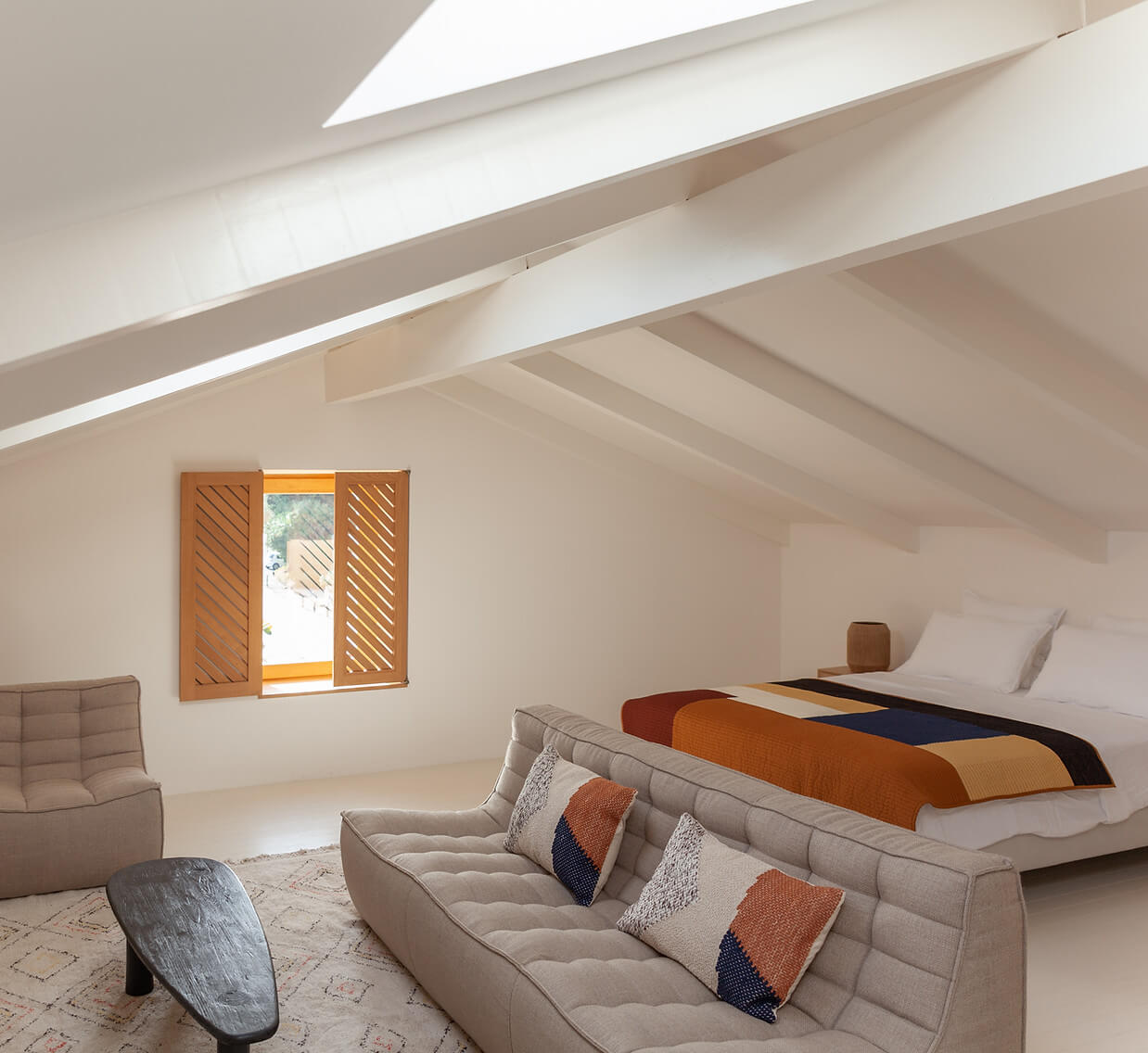 Casa Santa Teresa, Europe — Minimalist attic bedroom with exposed beams, skylight, linen sofa, warm textile accents, and views through wooden shutters.