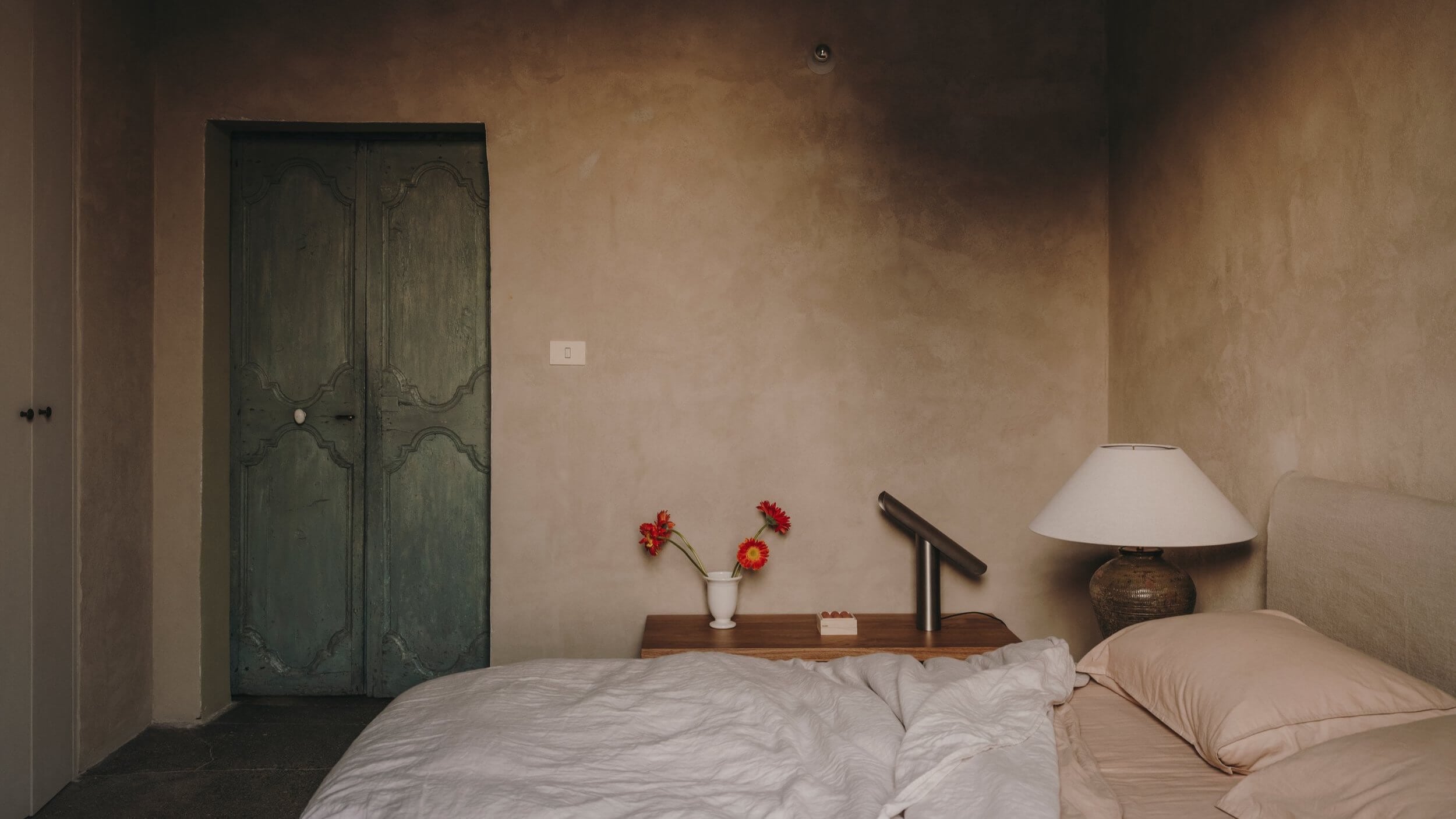 Casa Soleto, Europe — Minimalist bedroom with sage green paneled doors, neutral plaster walls, white bedding, wooden nightstand with red flowers and table lamp, morning light.