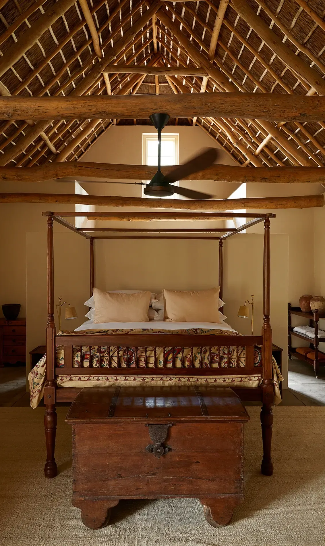 Sterrekopje, Africa — Four-poster wooden bed with cream linens beneath exposed timber roof beams and black ceiling fan in minimalist bedroom.