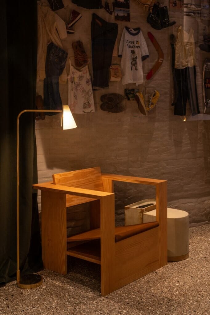The Venice Venice Hotel, Europe — Minimalist wooden chair beneath brass arc lamp beside gallery wall displaying vintage clothing and accessories.