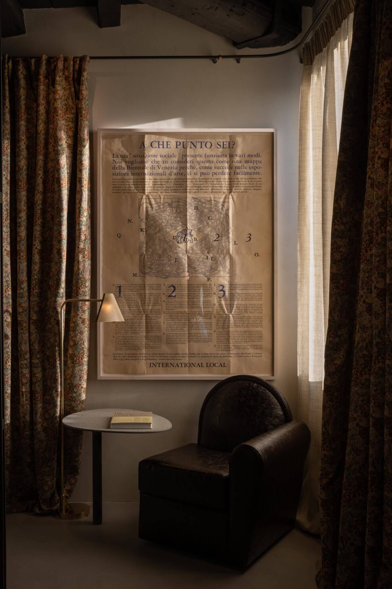 The Venice Venice Hotel, Venice, Italy — Gallery alcove with framed Italian exhibition poster, black leather chair, patterned curtains, and ambient reading light.