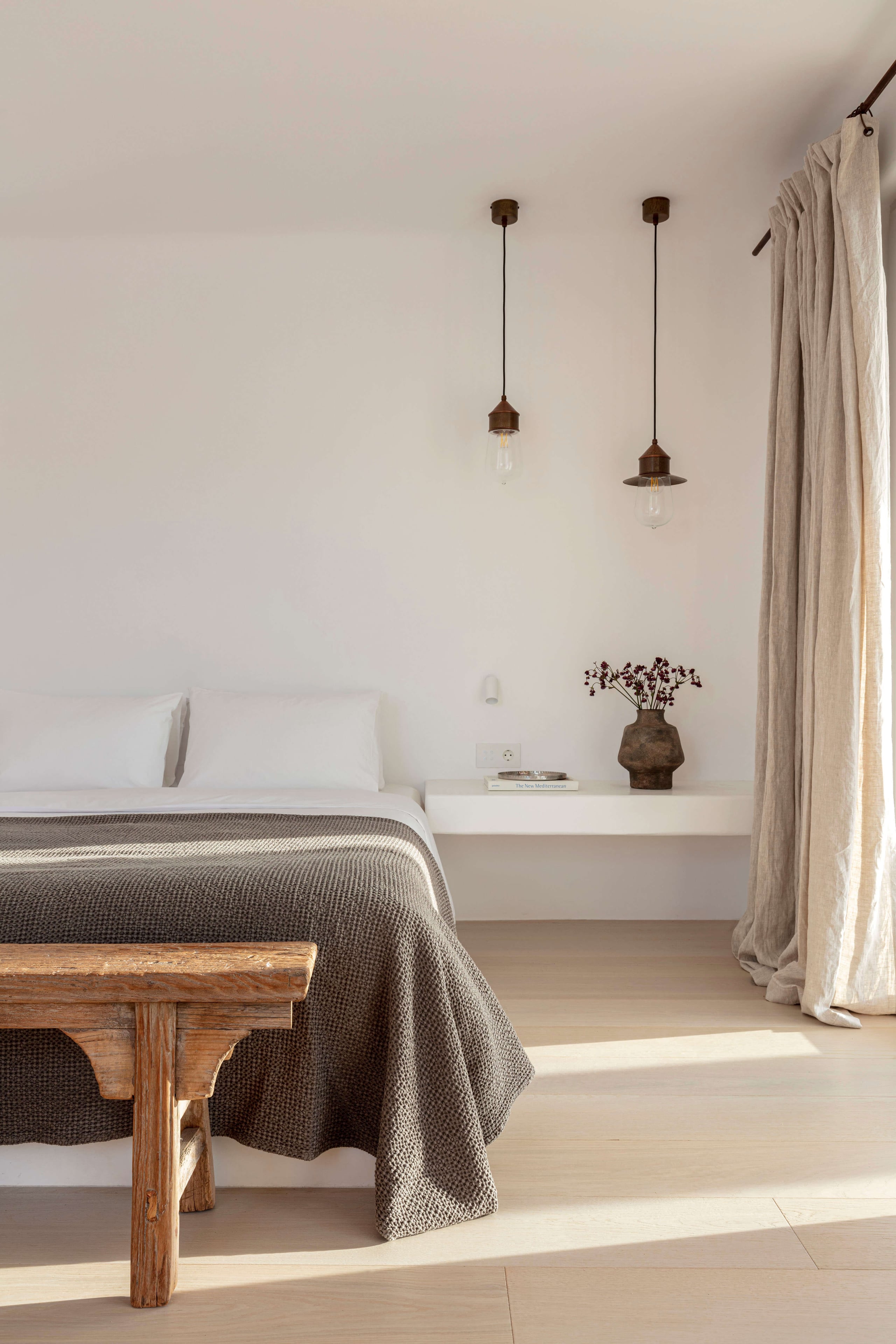 Villa Tama, Europe — Minimalist bedroom with suspended bronze pendant lights, linen curtains, wooden bench, and neutral palette bathed in natural light.