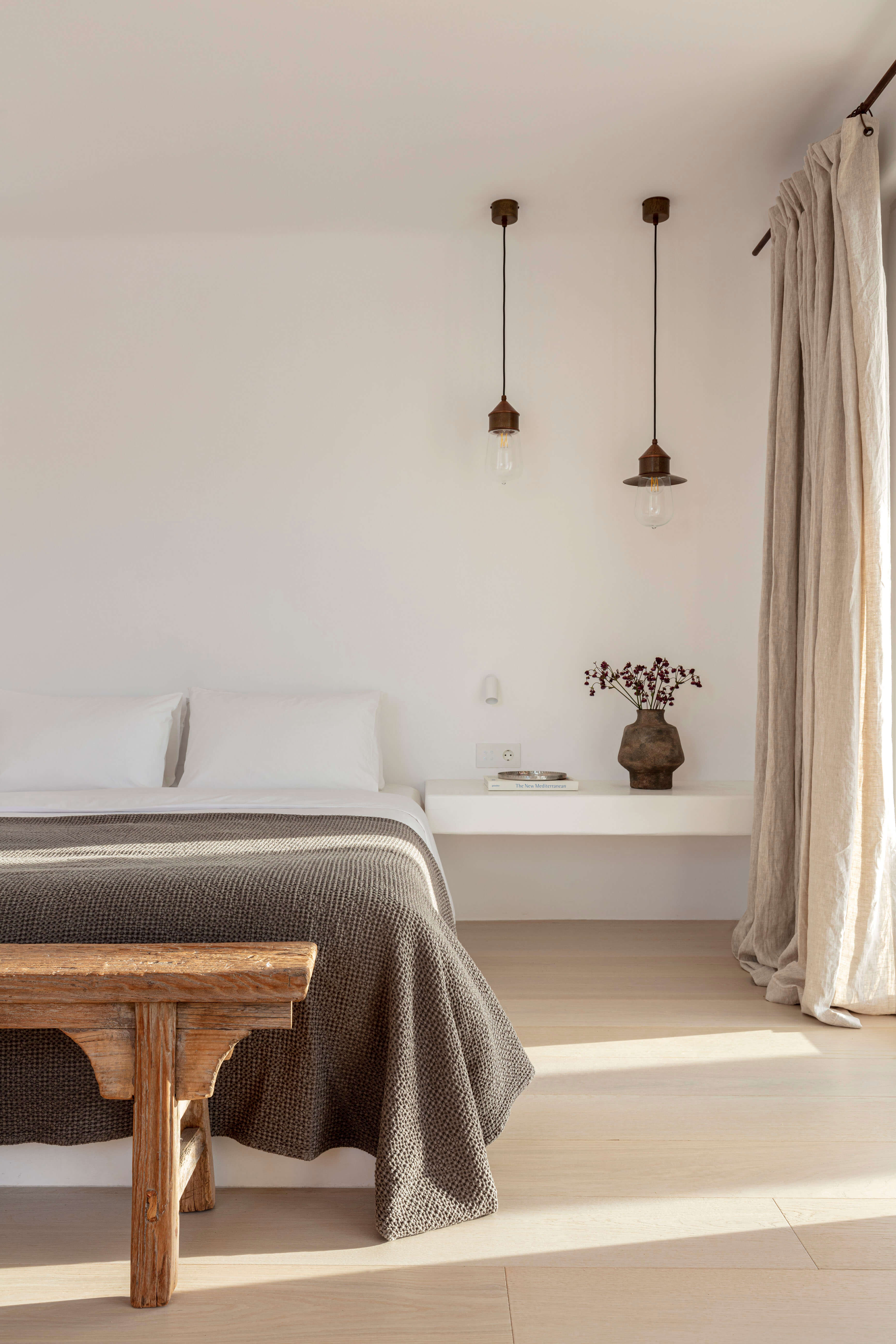 Villa Tama, Europe — Minimalist bedroom with suspended bronze pendant lights, linen curtains, wooden bench, and neutral palette bathed in natural light.