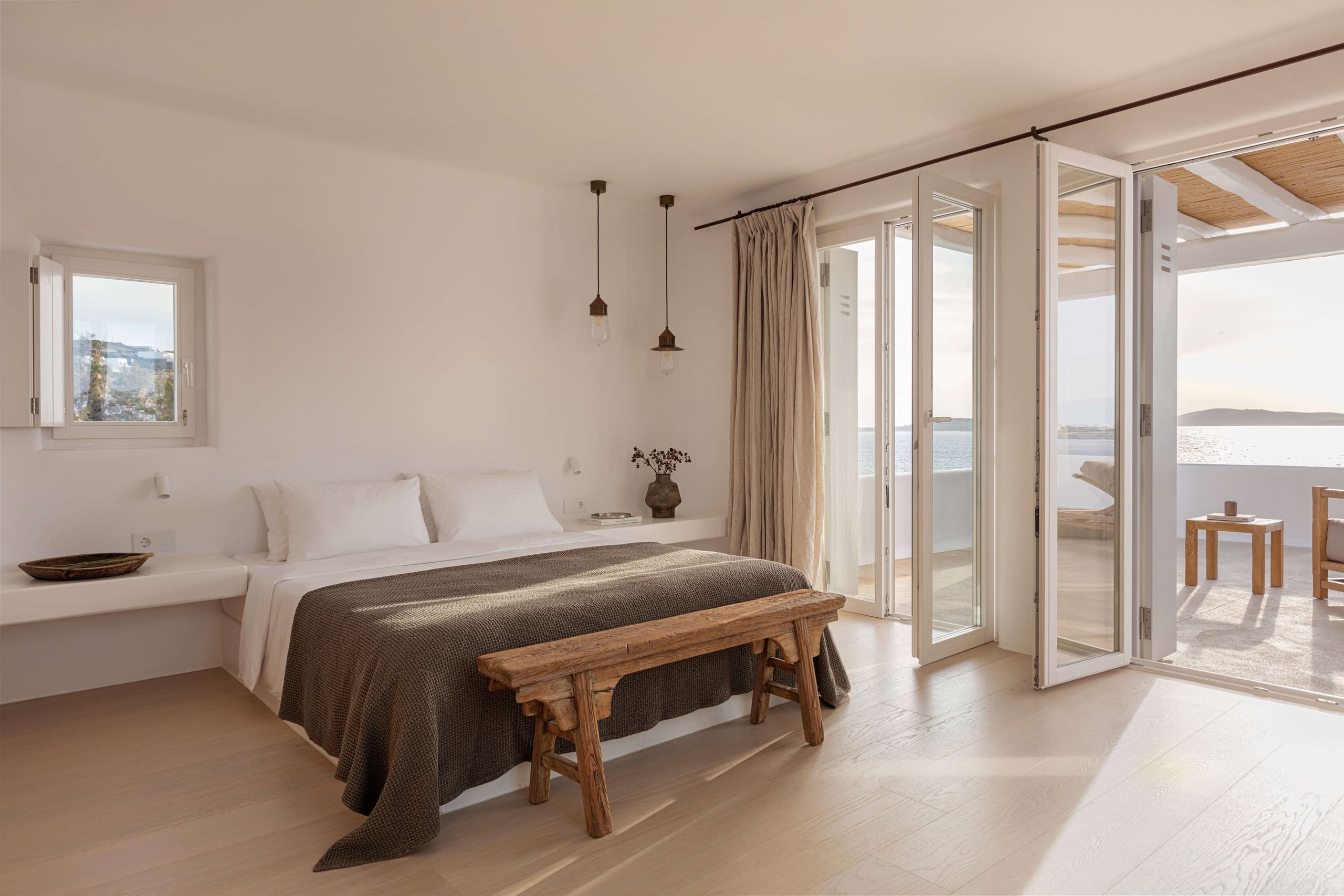 Villa Tama, Europe — Minimalist bedroom with sea-view terrace, wooden bench, pendant lights, soft linens, and floor-to-ceiling glass doors opening to Aegean landscape.