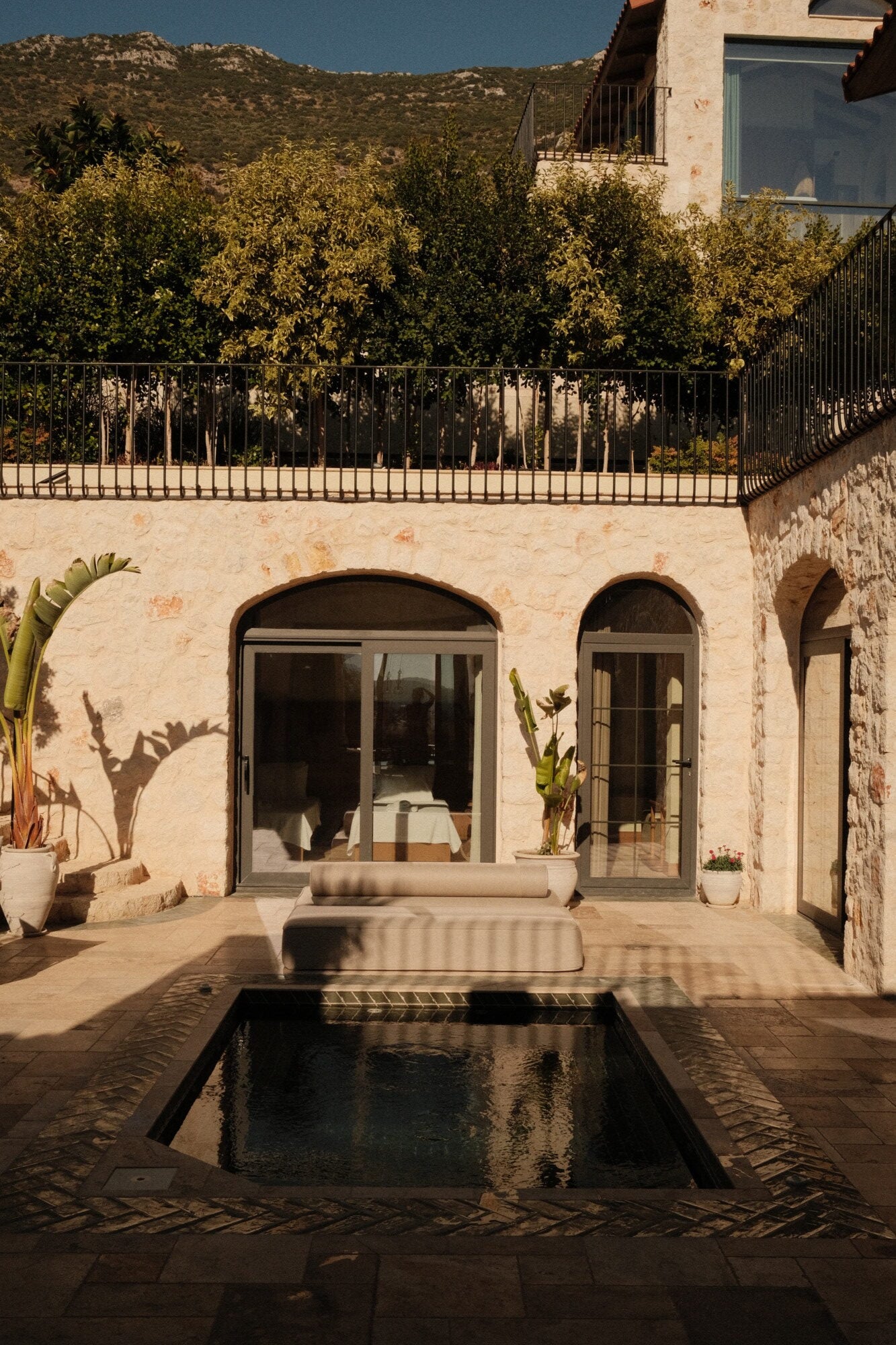 Villa Thalia by Muses Kalkan, Kalkan, Turkey — Sunlit courtyard with reflecting pool, arched stone doorways, and terraced gardens against hillside backdrop.