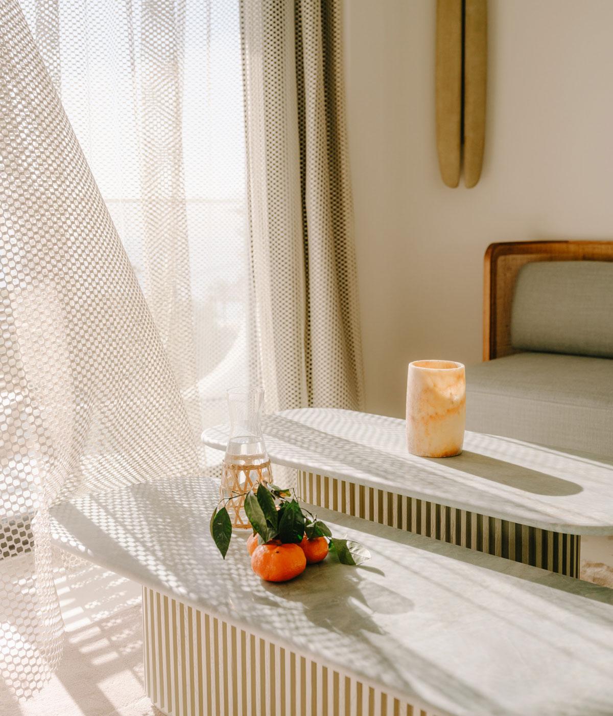 Belle Plage, Europe — Sunlit minimalist bedroom with marble surfaces, textured linen curtains, fresh citrus, and mid-century wooden furniture casting geometric shadows.