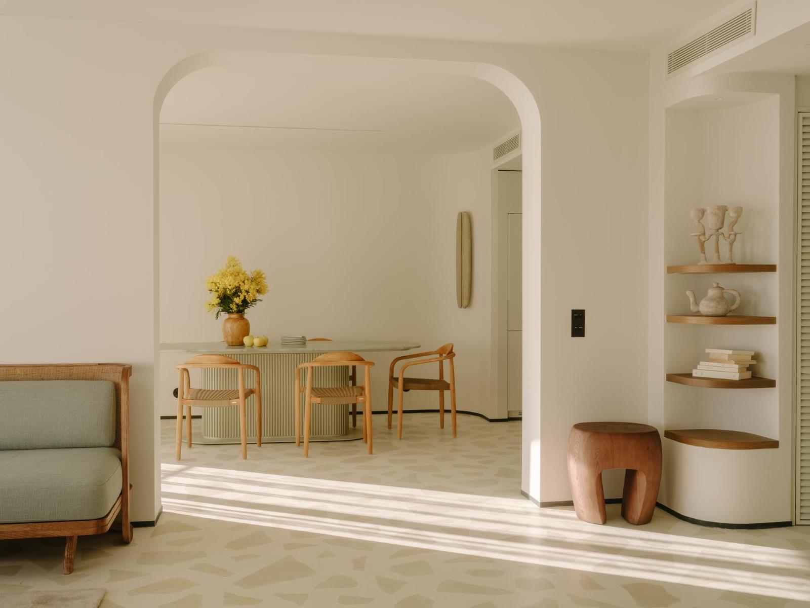 Belle Plage, Europe — Minimalist interior with rounded archway, wooden furniture, cream walls, floating shelves, terracotta stool, and filtered natural light.