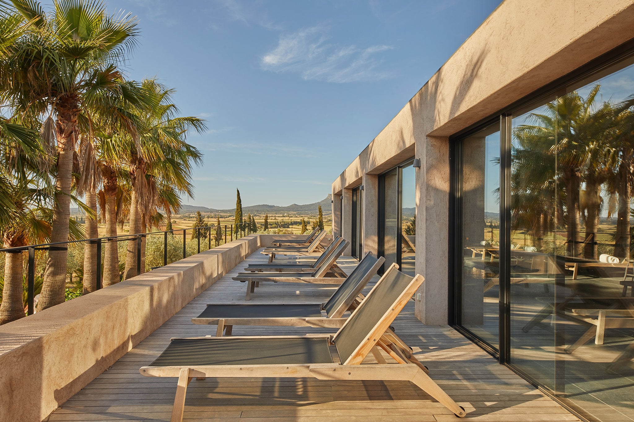 Finca Serena, Balearic Islands, Spain — Terracotta-walled modern pavilion with row of wooden loungers overlooking Mediterranean landscape, palm trees, and distant mountains in golden afternoon light.