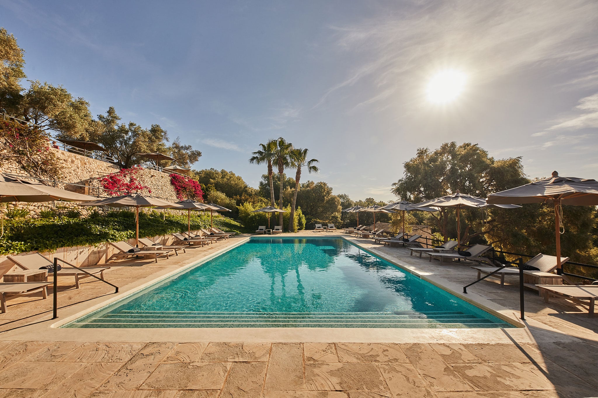 Finca Serena, Balearic Islands, Spain — rectangular lap pool with turquoise water, limestone terrace, lounge chairs under pergolas, blooming bougainvillea, palm trees, golden-hour sunlight.