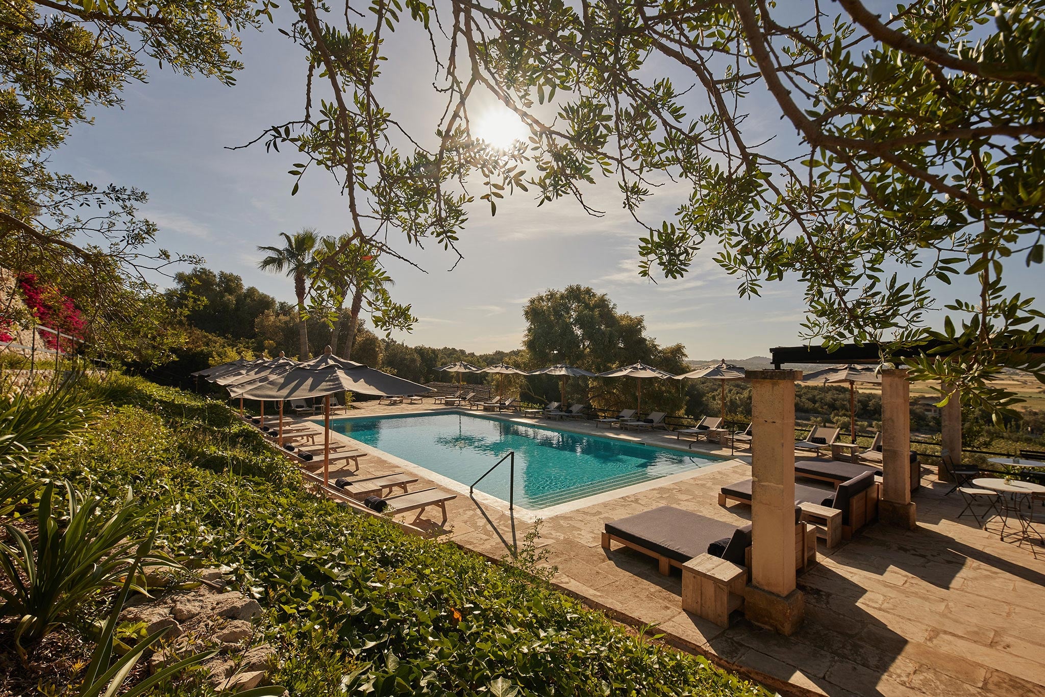 Finca Serena, Balearic Islands — Resort pool terrace with loungers, olive branches overhead, Mediterranean landscape, striped umbrellas, stone columns, midday light.