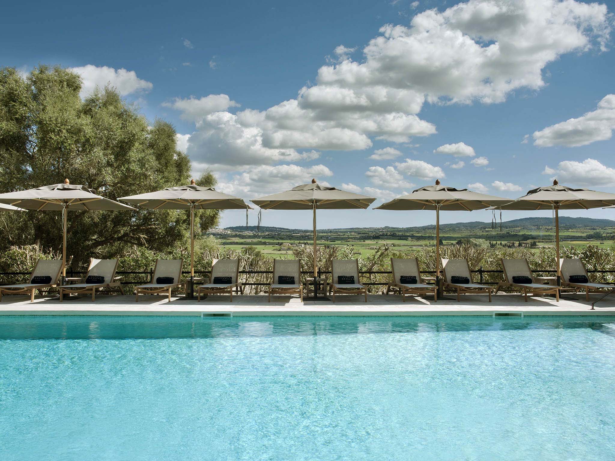 Finca Serena, Balearic Islands — Infinity pool with turquoise water, cream umbrellas, teak loungers, olive trees, and rolling countryside vista under blue sky.