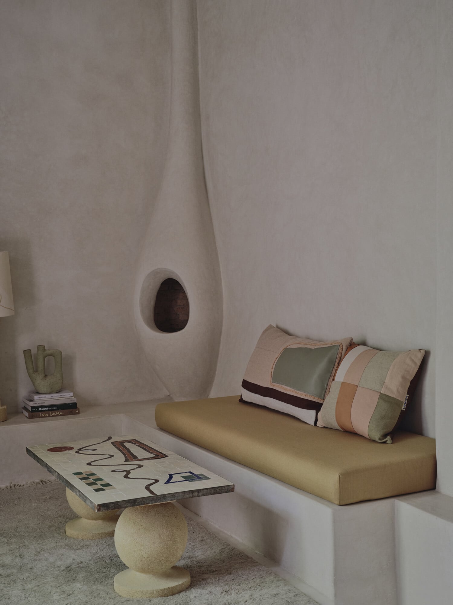 Rosemary, Africa — Minimalist whitewashed room with curved architectural niches, cream daybed, patterned pillows, sculptural stone side table, and abstract artwork.