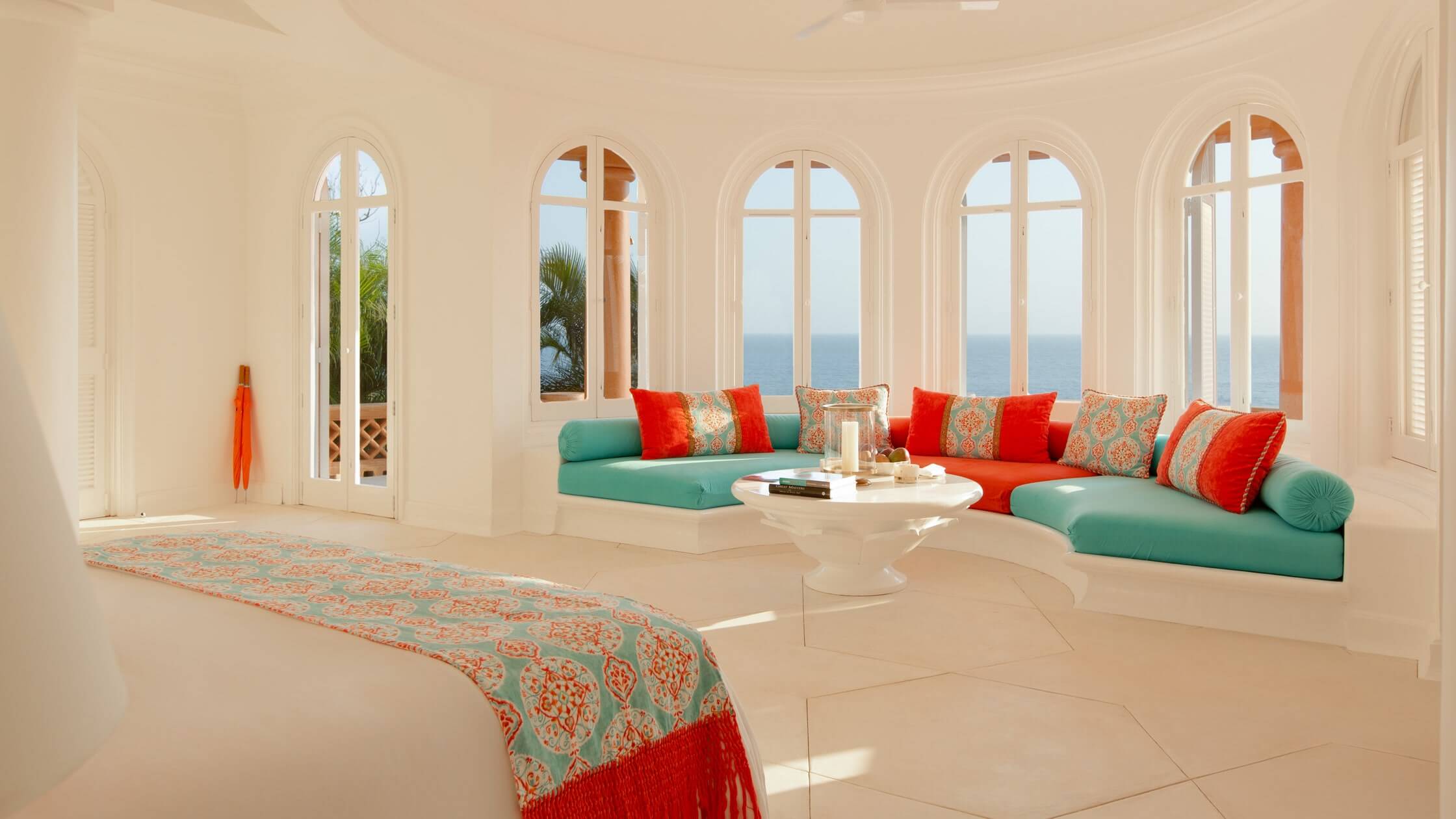 Cuixmala, Mexico — Coastal living room with turquoise and coral modular seating, arched windows overlooking ocean, travertine floors, patterned pillows, sculptural table.