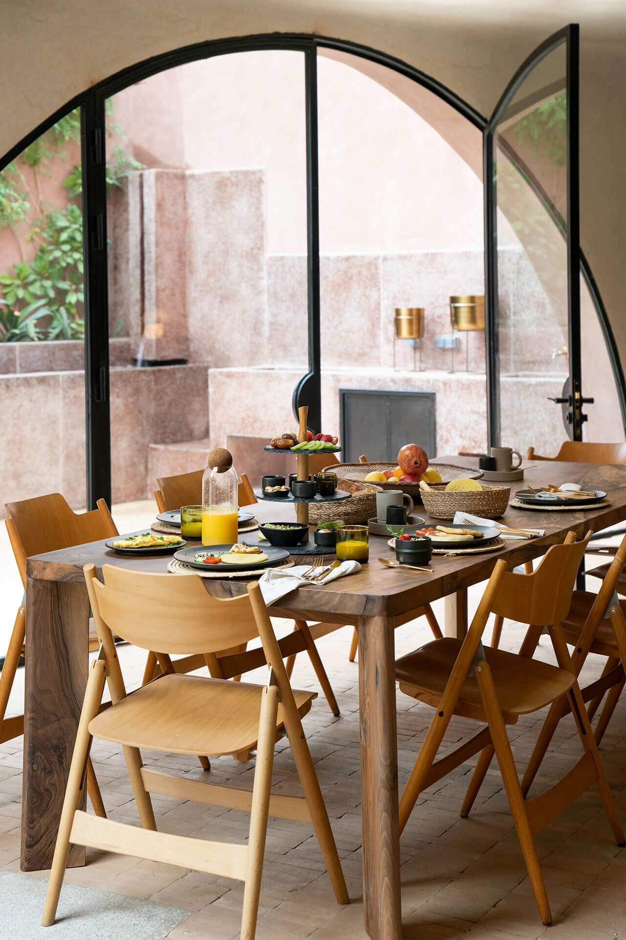 Maison Brummell Majorelle, Africa — Dining terrace with wooden table, mid-century chairs, arched black-framed windows overlooking courtyard, morning light, breakfast service setup.