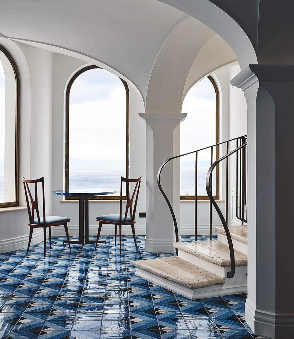Borgo Santandrea, Europe — Arched colonnade with geometric blue tilework, mid-century chairs, and Mediterranean sea views through tall windows.
