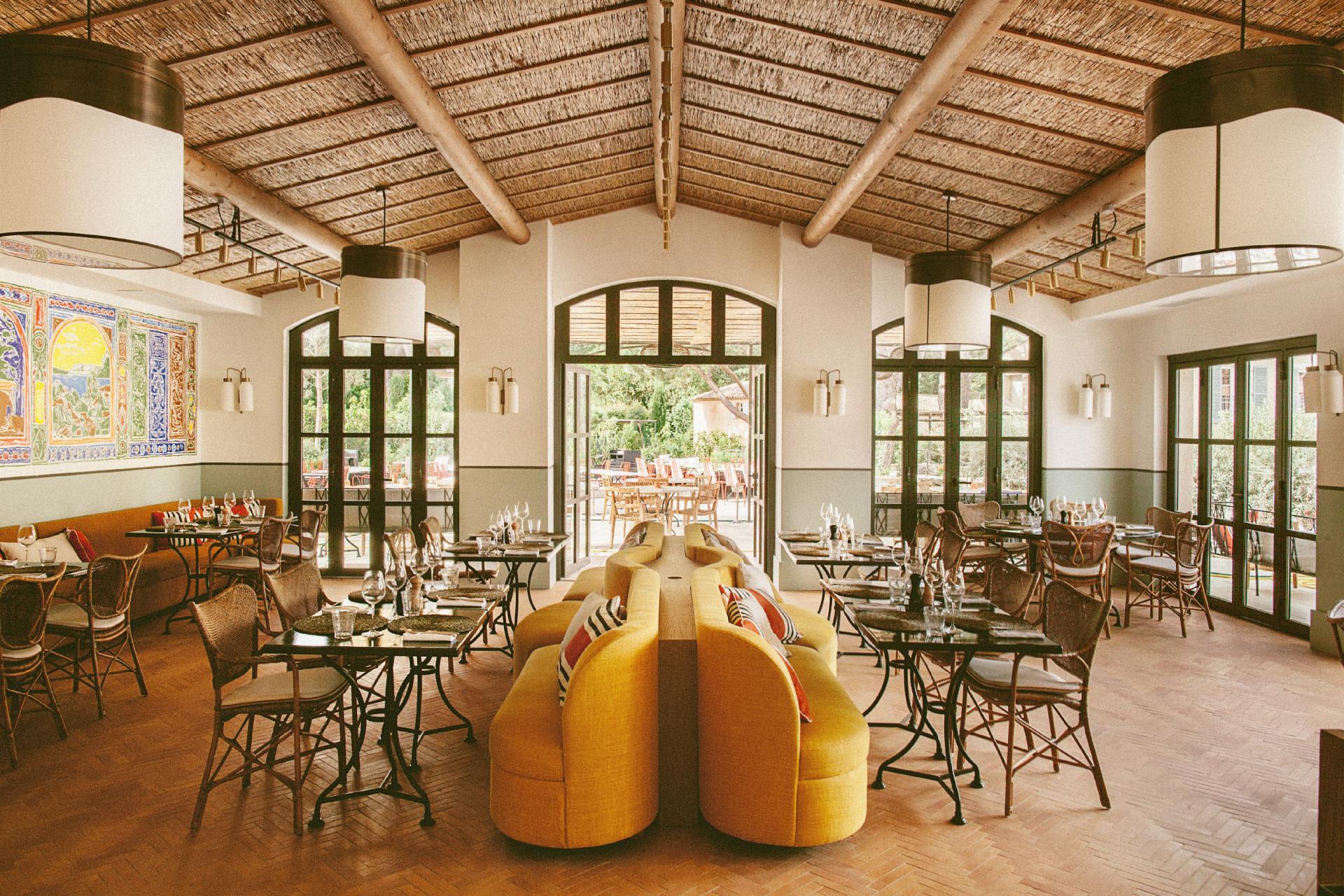 Hôtel Lou Pinet, Europe — Dining room with ochre curved banquette, rattan ceiling, arched windows, brass sconces, and terracotta tile floors opening to garden.