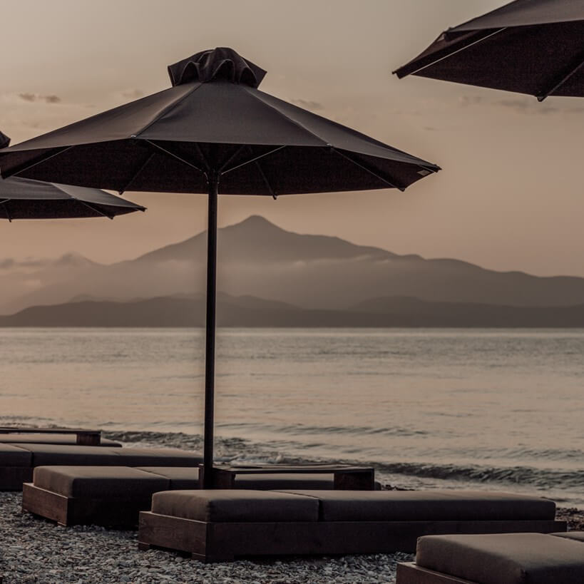 Casa Cook Samos, Greece — Black umbrellas and loungers on sandy beach at dusk with mountain silhouette across calm sea.