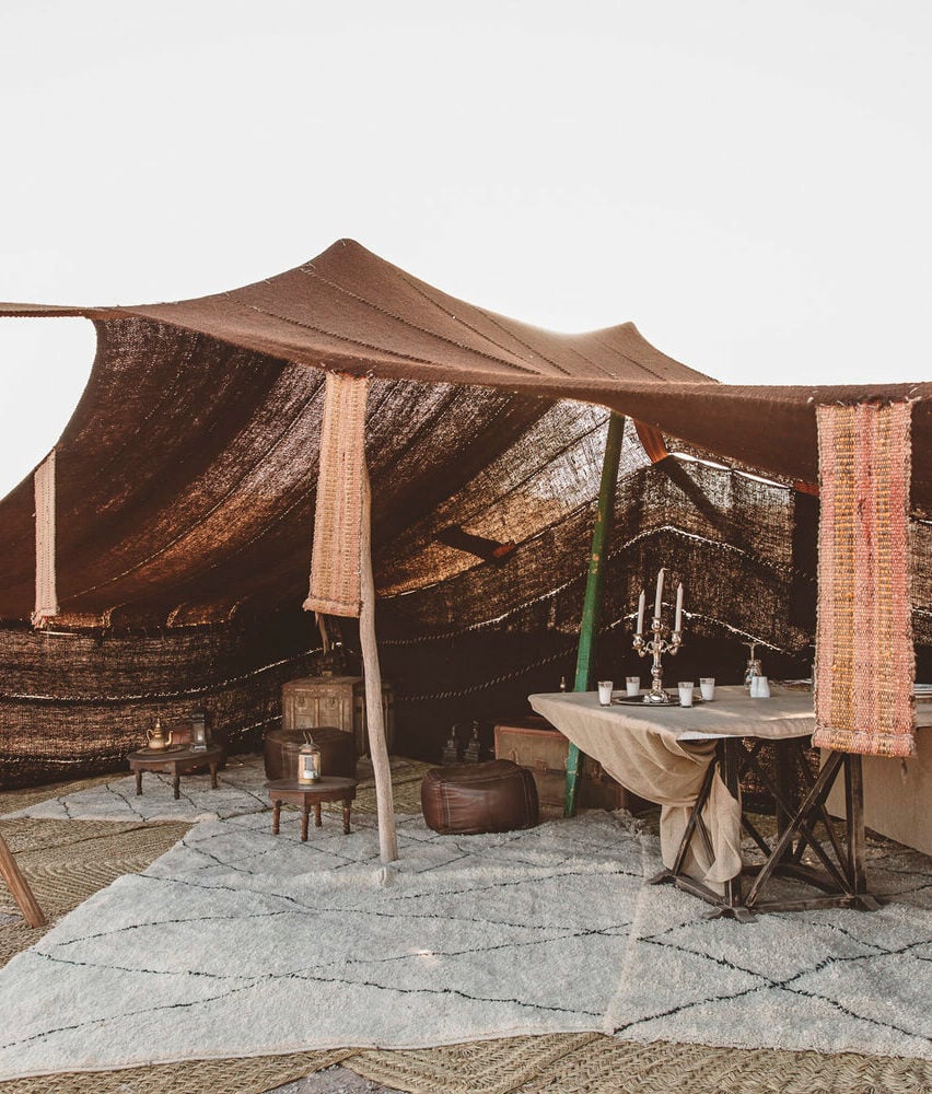 Scarabeo Camp, Africa — Draped canvas pavilion with brick pillars, rustic dining table, lanterns, and layered textile furnishings on stone paving in warm earth tones.