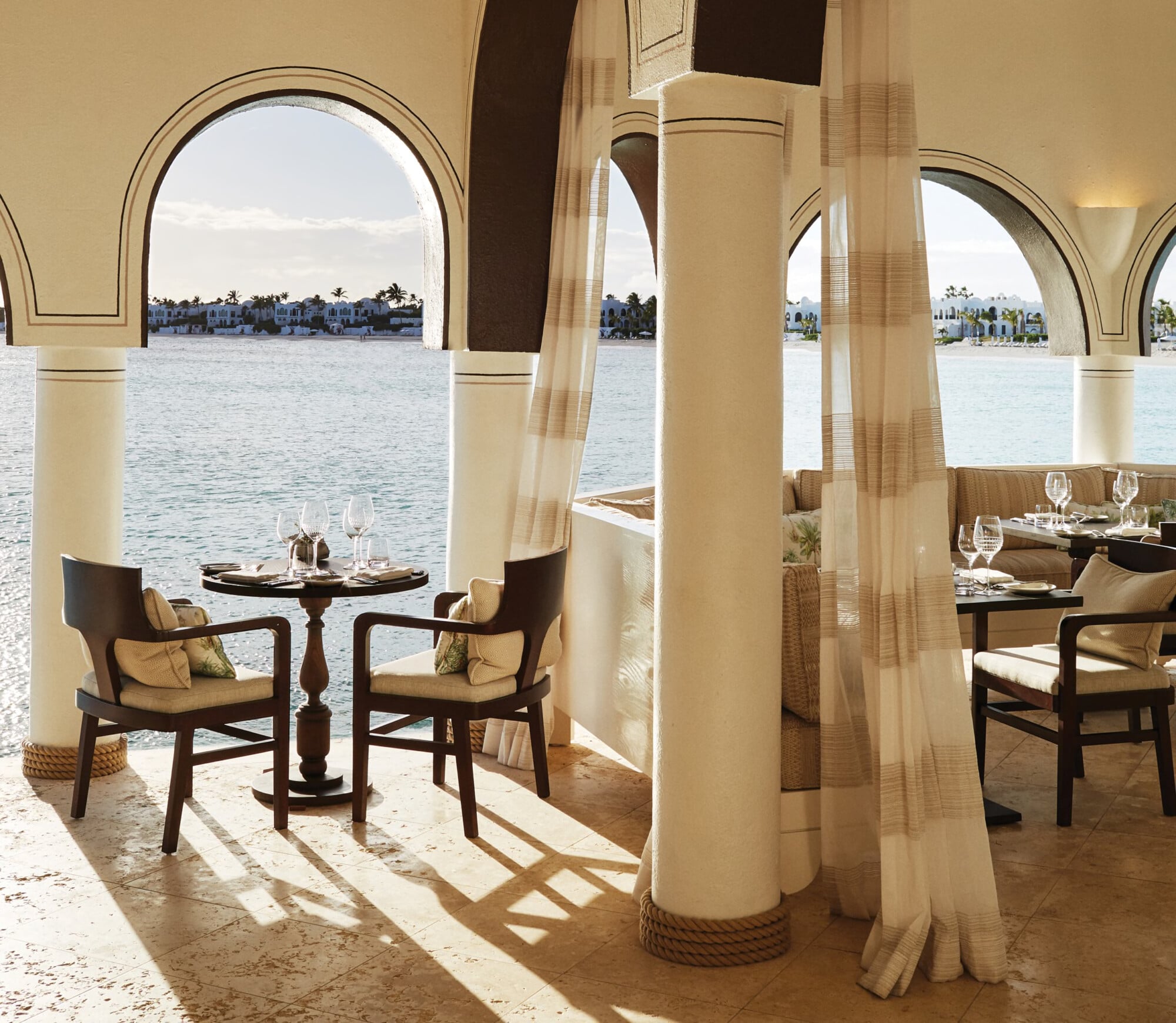 Pimms, North America — Beachfront dining pavilion with arched openings, sheer curtains, wooden furniture, and waterfront views of residential architecture.