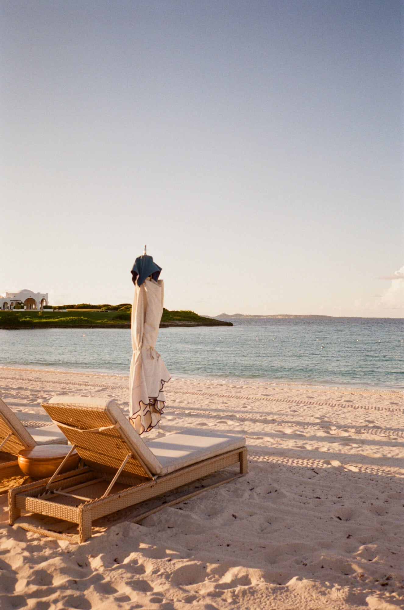Cap Juluca, Anguilla — Woven lounge chairs and cream umbrella on white sand beach at sunset, turquoise water and distant headland beyond.
